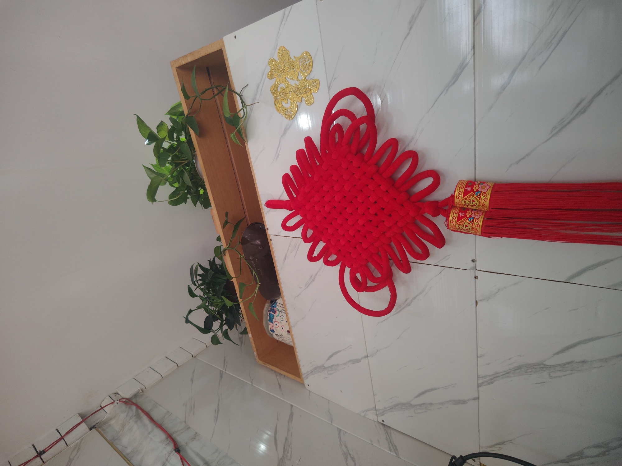 This is a ground-level, eye-level, medium shot of a wall decoration. The wall appears to be made of large, white marble-patterned tiles. A red, intricate knot ornament hangs from the wall. Above the knot, a wooden shelf holds two potted green plants, one with trailing vines, and some other objects. To the right of the knot, a gold, stylized Chinese character ornament is affixed to the wall. The scene is indoors, likely in a residential setting. There is no indication of Chongqing High-tech Zone, China, in the image.