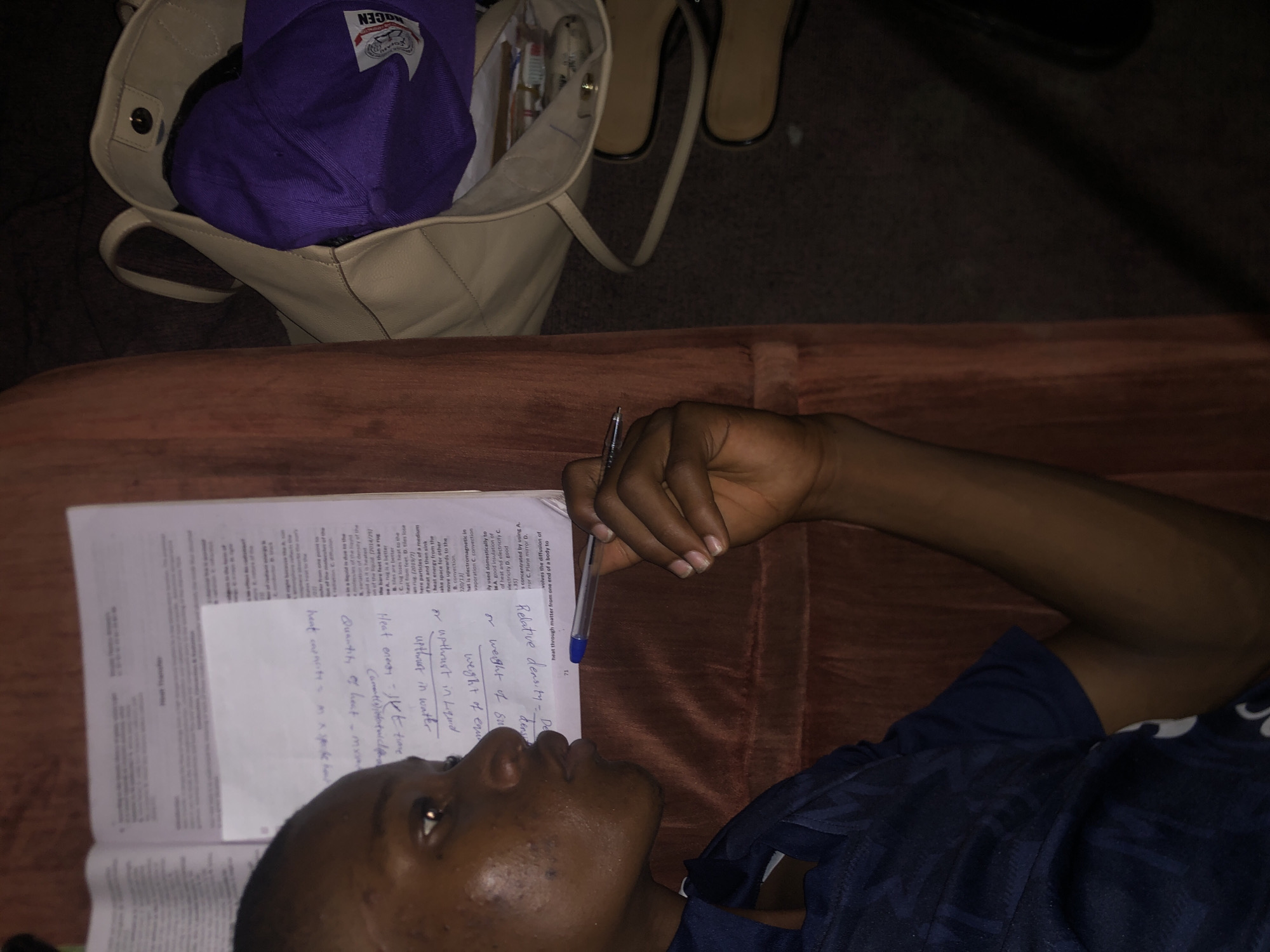 A young Black man is lying on a brown couch, possibly in a home in Atani, Nigeria. He is wearing a dark blue shirt. He is holding a blue pen in his right hand and appears to be studying a textbook and notes on a white sheet of paper. The text in the book and on the notes is partially legible. A beige purse with a purple hat inside is visible near the top left of the image. Sandals are near the purse. The lighting is dim.