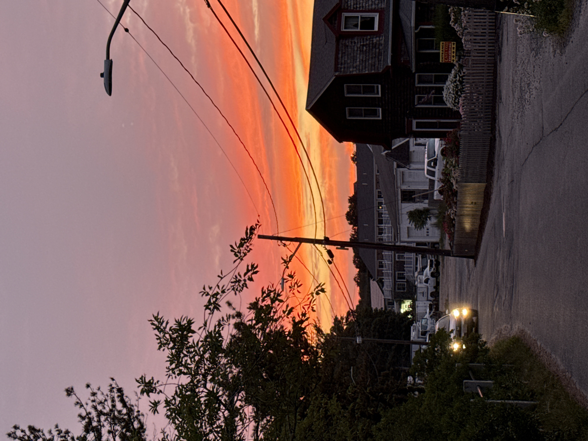 The image depicts a street scene at dusk in Provincetown, Massachusetts. The sky is illuminated with a vibrant gradient of pink and orange hues, indicative of a sunset.
In the foreground, a paved road curves to the left, bordered by lush green foliage on the left and a wooden picket fence on the right. A car with its headlights on is visible driving on the road. On the right side of the road, a dark-colored, multi-story building with shingled siding and dormer windows is prominent. A yellow sign with red lettering is affixed to the fence in front of this building, partially obscured by foliage.
Further down the road, several other buildings, including what appears to be a larger structure with multiple windows and balconies, are visible. A street lamp is positioned in the upper right quadrant of the image. Utility wires are strung across the sky. Various vehicles, including trucks and what looks like a recreational vehicle, are parked or driving in the background. The overall impression is a coastal town street bathed in the warm light of twilight.
