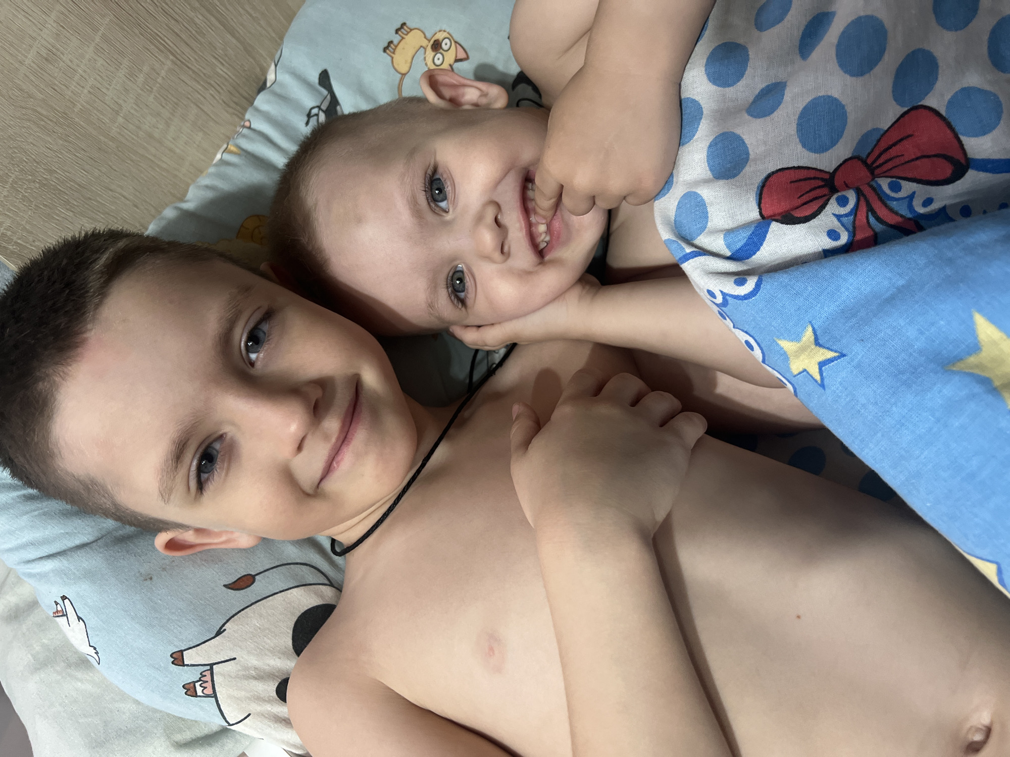 Two fair-skinned children lie on a patterned pillow and under a blue and white patterned blanket. The older child has short dark hair and is wearing a black necklace. The younger child has fair hair and is grinning, with their fingers in their mouth. The pillow has cartoon images of animals. The blanket is blue with yellow stars and has a white section with blue polka dots and red bows. The location is Марковский, Russia.