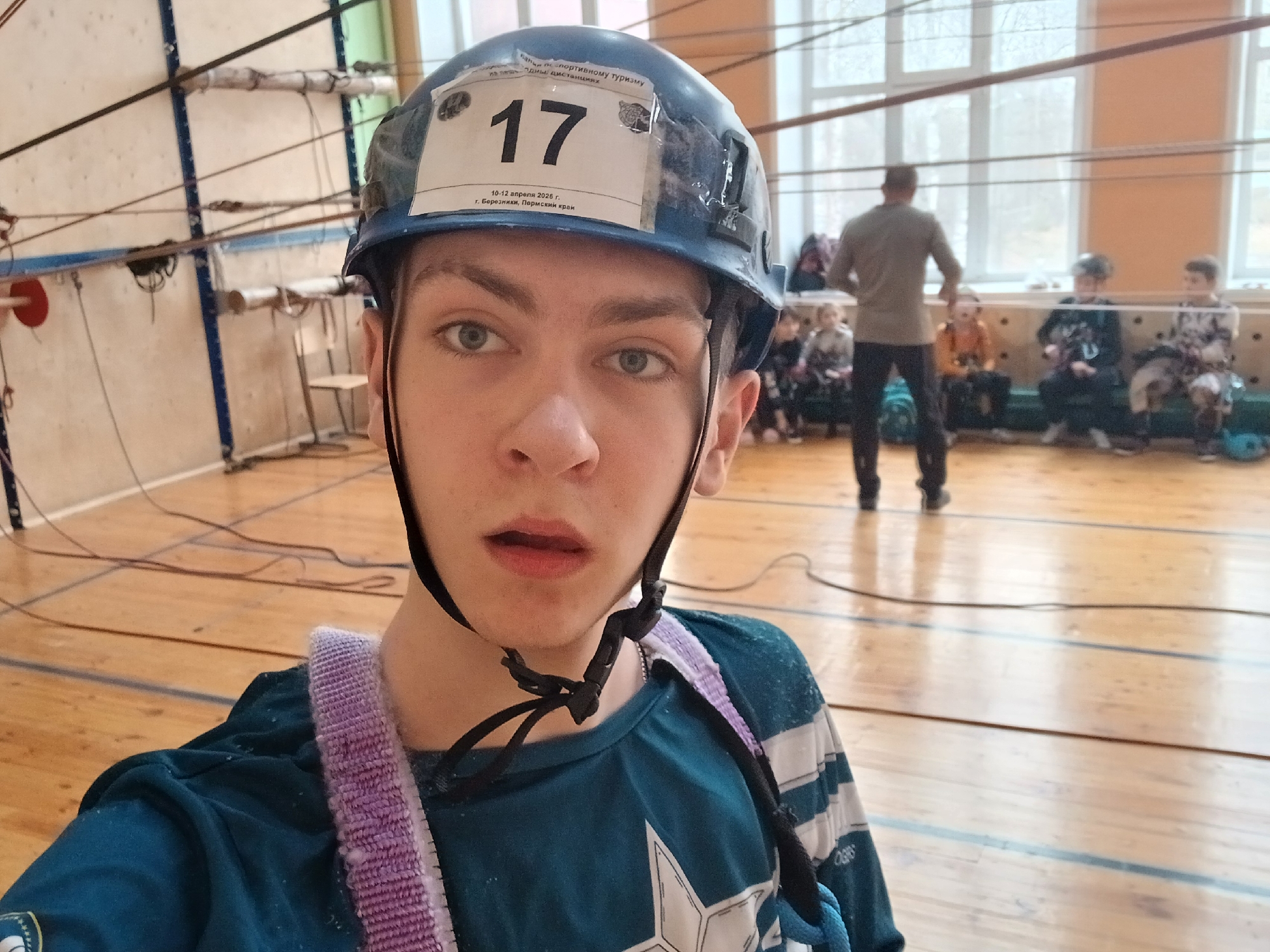 A young person is taking a selfie in what appears to be a gymnasium. They are wearing a blue helmet with the number "17" attached to the front, along with some text indicating a sports tourism event held in Berezniki, Perm Krai, Russia, between April 10-12, 2026. The person is wearing a blue and white striped shirt with a white star and a climbing harness with a purple strap. The background shows a wooden gym floor, climbing ropes, a window, and a group of people sitting and standing, with a man in the foreground facing away from the camera.