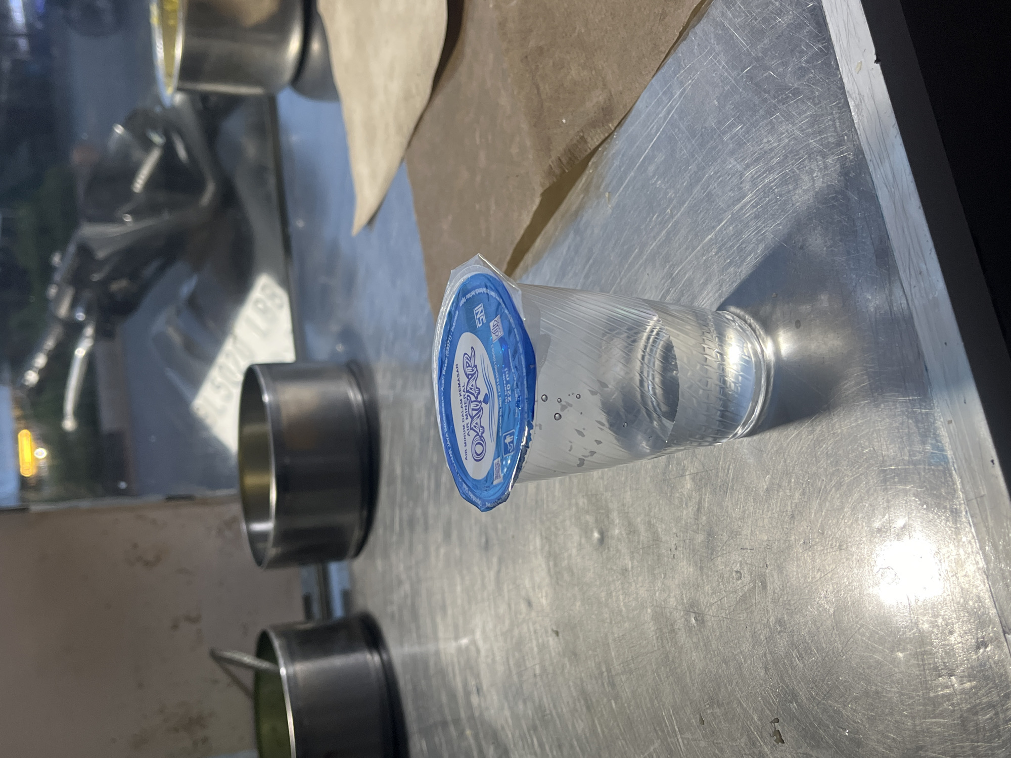 A plastic cup filled with clear liquid and sealed with a blue lid sits on a metallic surface. The lid has blue text and a logo that appears to say "OASIS AIR MINERAL". The cup has condensation on the outside and small bubbles are visible inside the liquid. In the background, there are two metallic bowls and a motorcycle is partially visible through a window. The scene appears to be outdoors, possibly at a street food stall, in Banda Aceh, Indonesia.