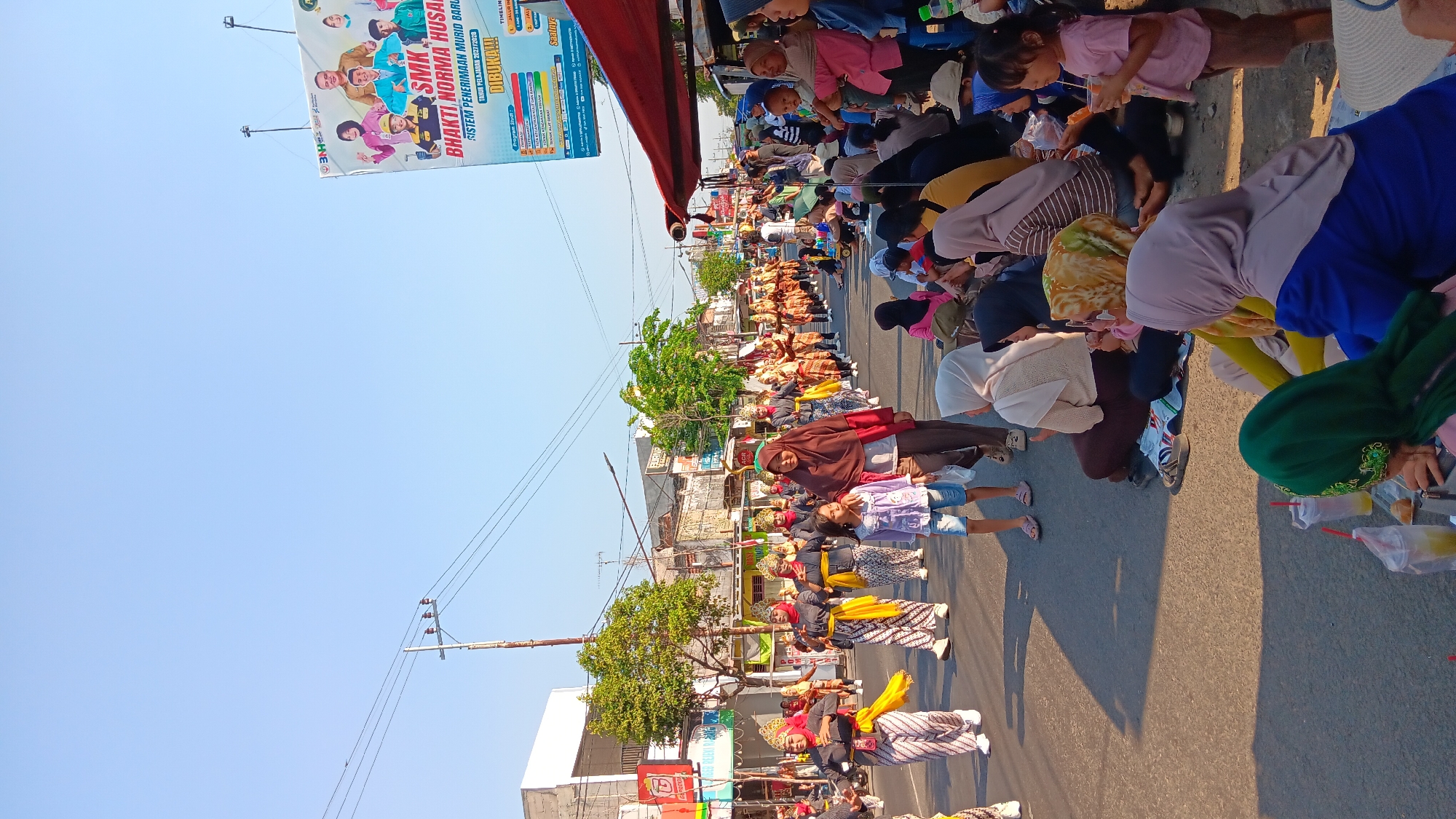 The image depicts a street scene in Tanjung Anom, Indonesia, featuring a parade and spectators. In the foreground, a crowd of people, predominantly women and children, are seated on the ground, observing the procession. They are dressed in various casual and traditional attire, with many wearing hijabs. Some individuals are holding drinks or food items.
The parade participants, dressed in traditional costumes, are walking down the street. The costumes appear to be colorful and elaborate, suggesting a cultural or festive event. The parade route is on an asphalt road, with buildings and trees lining the sides. A large billboard for "SMK BHAKTI NORMA HUSADA" is visible in the background, indicating the presence of a school or educational institution in the area. Power lines and utility poles are also present, characteristic of a street in a city or town. The sky is clear and blue, suggesting a sunny day. The overall atmosphere appears to be lively and community-oriented.