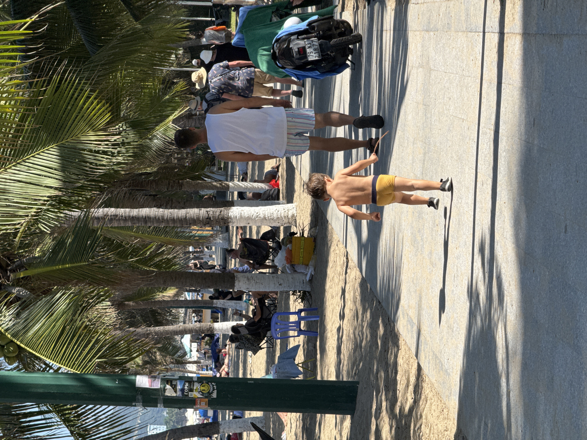 The image displays a sunny beachside scene along a paved promenade in Nha Trang, Vietnam. A sandy area borders the paved walkway to the left and partially to the right.
In the foreground, a young child, bare-chested and wearing yellow briefs and dark athletic shoes, walks away from the camera on the paved path, holding a short stick in their right hand. Directly ahead, an adult male, wearing a white tank top, vertically striped swim shorts, and dark flip-flops, also walks away from the camera on the same path. Further down the path, several other adults are observed walking, some wearing hats.
To the immediate right of the paved path, a dark-colored scooter or motorcycle is parked, partially covered by a green tarp and a blue towel. A Vietnamese license plate, "79-M3 218.77," is clearly visible on the rear of the vehicle. A yellow box sits nearby.
The sandy area to the left of the walkway is populated by numerous tall palm trees with green fronds, some bearing coconuts. Their trunks vary in appearance, with some painted white at the base. Under the shade of these trees, multiple individuals are present; some are seated on black folding chairs, one person is lying on a red towel in the sand, and others are standing or sitting informally. A single blue plastic chair and a small yellow table are visible in this area. A prominent dark green utility pole stands in the left foreground, adorned with several layered stickers, including text in Cyrillic ("ГОРЫ" - Mountains) and a yellow smiley face emoji.
In the distant background, hills or mountains are visible beneath a clear sky. The scene is illuminated by bright sunlight, casting sharp, elongated shadows from the palm fronds and figures onto the paved walkway and sand.