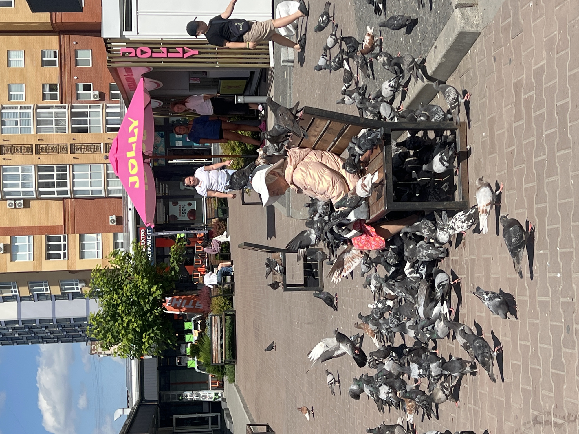 A bright, sunny day is captured in this outdoor scene, likely in the city of Ternopil, Ukraine, given the presence of a Jolly umbrella and nearby buildings. In the foreground, a woman wearing a light-colored jacket and a wide-brimmed hat is seated on a wooden bench, surrounded by a large flock of pigeons. Some of the pigeons are perched on the bench with her, while others are gathered on the brick-paved ground, actively feeding. The woman appears to be interacting with the birds, though the exact nature of the interaction is unclear.
Behind the woman, several other people are visible. To the right, a young boy in a black t-shirt and beige shorts walks by, carrying a crossbody bag. Further back, three women stand under a bright pink Jolly umbrella, which is a prominent feature of the scene, suggesting a commercial establishment. One woman is wearing a white t-shirt, another a blue t-shirt, and the third a light pink t-shirt. They appear to be customers or staff.
The background is dominated by a multi-story building with a modern, somewhat segmented architectural style, featuring a mix of yellow and brick-colored facades. A tall, slender building with a darker facade and numerous windows is also visible to the left, partially obscured by a lush green tree. Several other smaller structures and market stalls are present, with signage visible, including what appears to be a STIHL advertisement. The ground is paved with reddish-brown bricks, and a low concrete curb runs along the right edge of the frame. The overall impression is a busy urban public space, possibly a market square or pedestrian area, with a focus on the interaction between people and wildlife.