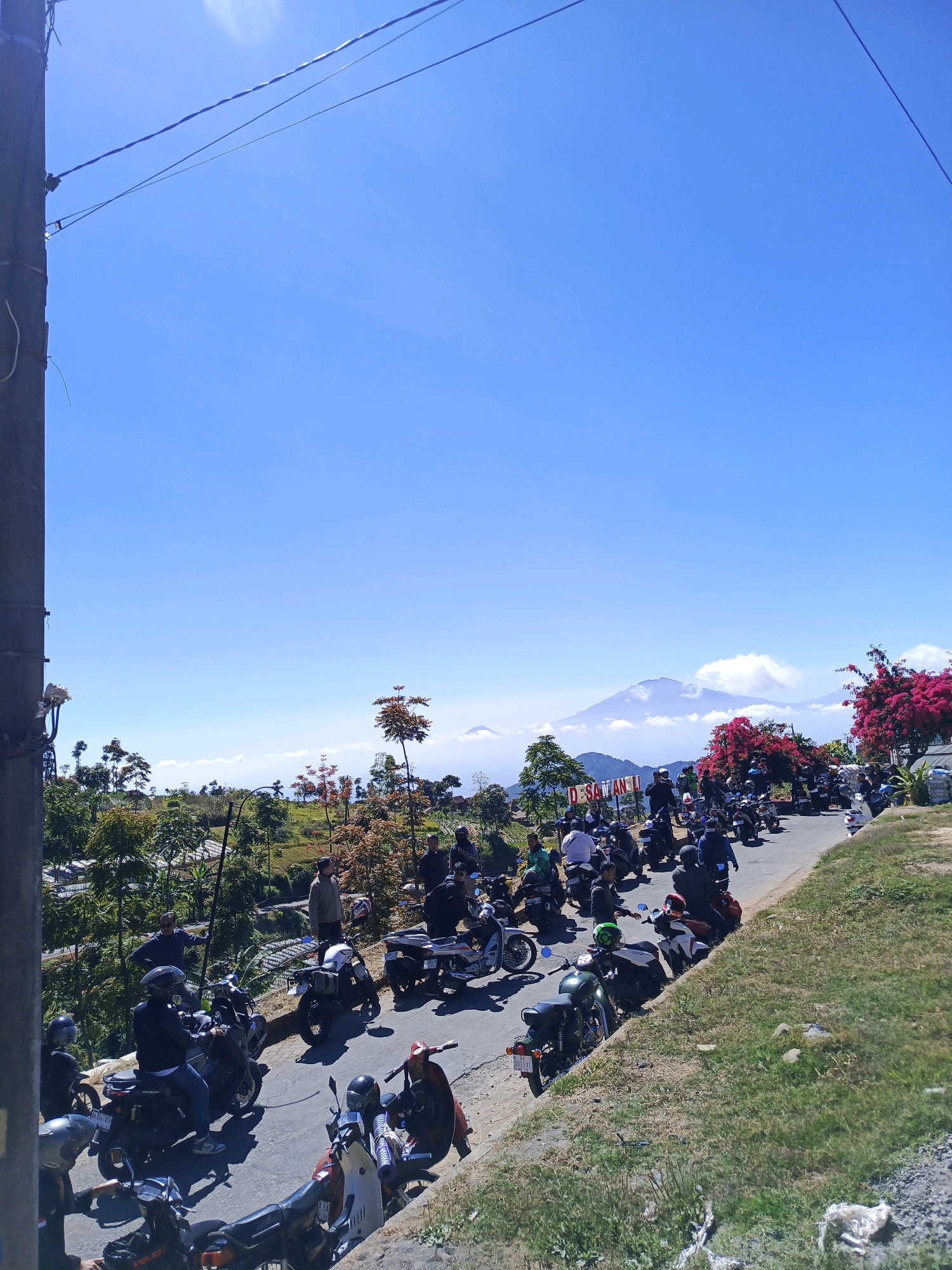 In Mangli, Indonesia, a gathering of people on motorcycles lines a paved road with a steep, grassy embankment on the right. The road winds uphill, with a dense green landscape of trees and foliage on the left. In the distance, a majestic mountain range, partially shrouded in clouds, forms the backdrop under a clear, bright blue sky. A red and white sign with the text "DESA TANGLI" is visible on the left side of the road. The scene appears to be a scenic viewpoint, possibly a tourist destination, with numerous motorcycles parked and people milling about. The overall impression is one of a lively, outdoor gathering in a beautiful, natural setting.