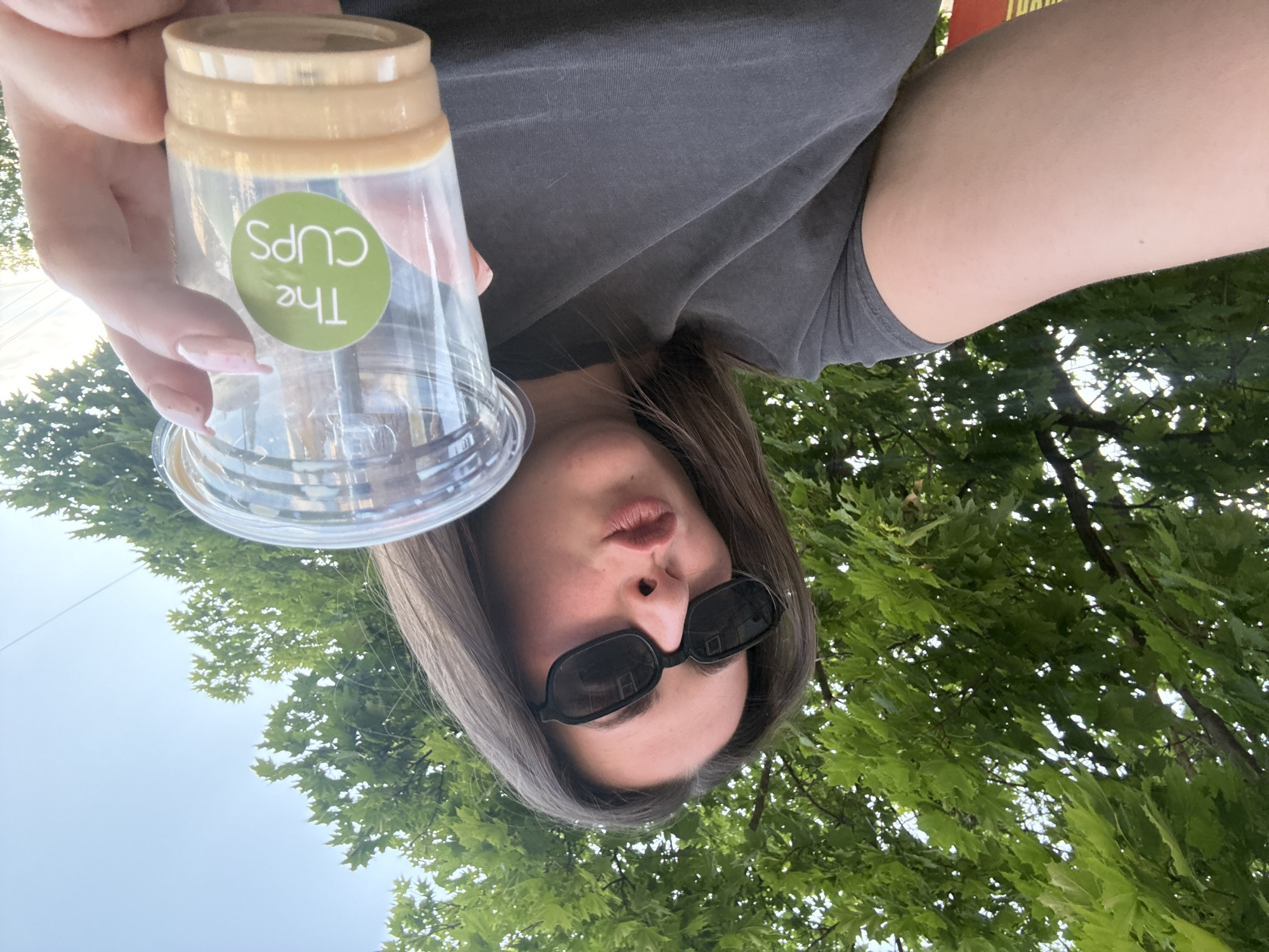A young woman with dark hair and black sunglasses is holding a clear plastic cup with a light brown beverage in it, and a green circular logo that reads "The CUPS". She is wearing a dark grey t-shirt and is outdoors, with green trees and a light blue sky in the background. She is looking towards the camera with her lips pursed.
