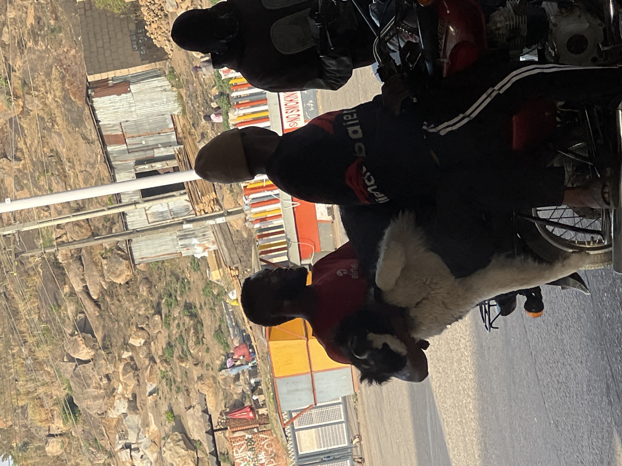 An eye-level, landscape-oriented photograph depicts a street scene in Jos, Nigeria, featuring several individuals and a dog around a parked motorcycle. The image, originally rotated 90 degrees clockwise, shows a sunny day with clear skies.
In the foreground and midground, a dark-colored motorcycle is central. A person wearing a dark jacket with white stripes (resembling Adidas branding) is seated on the motorcycle, with their foot resting near the pedal. Another individual stands directly beside the motorcycle, wearing a dark beanie/cap and a dark long-sleeved shirt that displays "BAILA" in white text and some red graphics. To their right, a younger person in a red short-sleeved shirt with the "Airtel" logo printed on it stands near a black and white dog with thick fur, which is perched comfortably on or next to the motorcycle.
The background reveals a street or dirt road leading to an area with various structures. These include corrugated metal shacks, some brick buildings, and colorful market stalls or goods. Further in the distance, a rugged, rocky, and hilly landscape with sparse vegetation is visible, consistent with the geography of Jos. Utility poles and overhead lines crisscross the scene.
A notable detail is a white sign with a red border that clearly reads "NO SMOKING." The overall impression is that of a casual, everyday interaction in a semi-urban or rural environment.