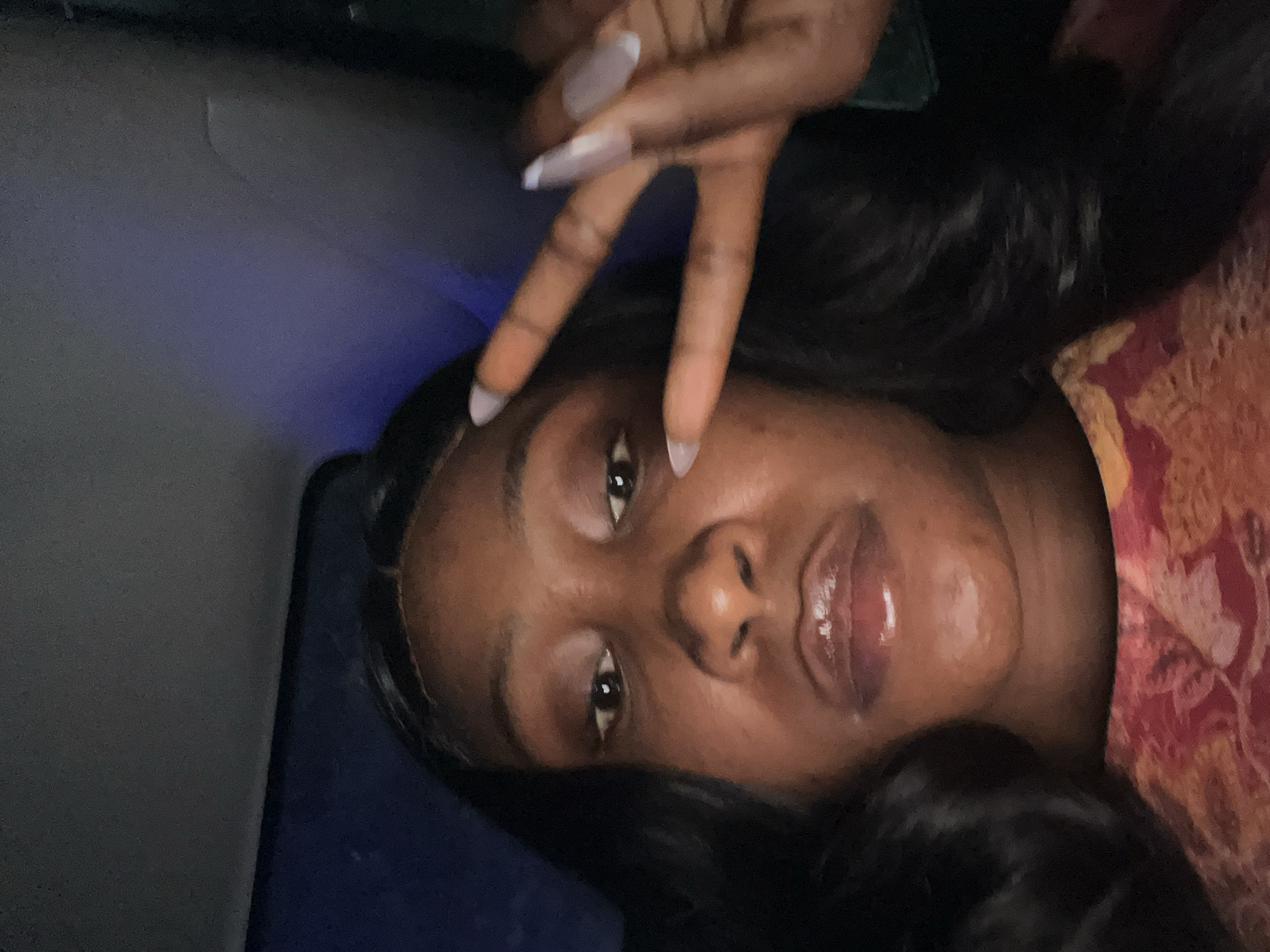A close-up, low-angle selfie shows a Black woman in what appears to be the interior of a vehicle, possibly at night. She is looking directly at the camera, with her face filling most of the frame. Her eyes are open, and she is wearing lip gloss. Her right hand is raised, with her index and middle fingers extended in a peace sign. Her fingernails are long and light-colored. She has dark, wavy hair. She is wearing a garment with a red and orange floral pattern. The background is dark and appears to be the interior of a car, with a dark ceiling and a blurred blue light source visible to the upper right. The image does not provide any specific visual cues that would allow for its location to be determined within Abuja, Nigeria.
