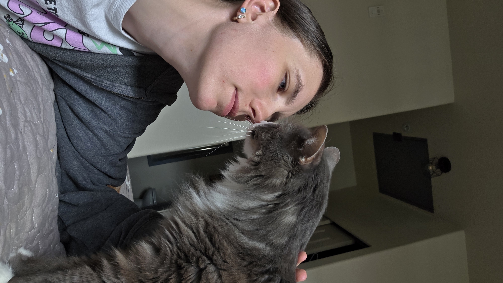 A young person and a gray cat are interacting on a bed, as suggested by the quilted bedding. The person, viewed from the side, is leaning in to nuzzle the cat's nose. Their eyes are closed, and they appear to be smiling, conveying a sense of affection. The cat, facing the person, has its eyes open and its nose touching the person's. The person is wearing a gray zip-up hoodie over a white t-shirt with a graphic design featuring a cartoonish character in pink and green. They also have a small, sparkling earring in their left ear. The background is dimly lit and shows a wall and a doorway with a light fixture visible. The scene suggests a moment of quiet intimacy and connection between the person and their pet. The time of day is not specified, but the indoor setting and lighting imply it could be anytime. The location is Beaverton, United States of America.