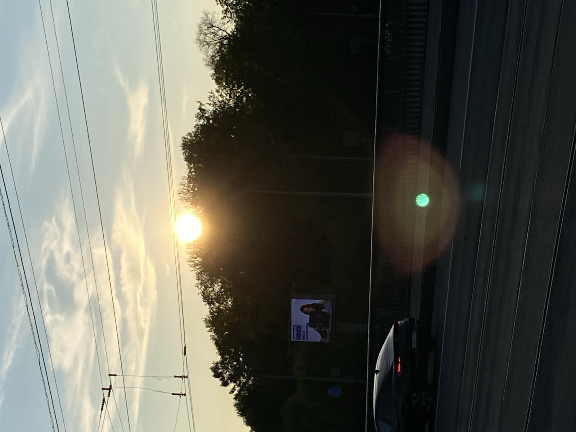 A sun shines brightly through trees, with utility poles and wires extending to the left. Adjacent is a multilane road, slightly curved and with a visible car on the right side of the frame. In the middle ground, a billboard features a woman. Given the visual cues (architecture, billboards, and trams), it is strongly likely this picture was taken in Saint Petersburg, Russia.