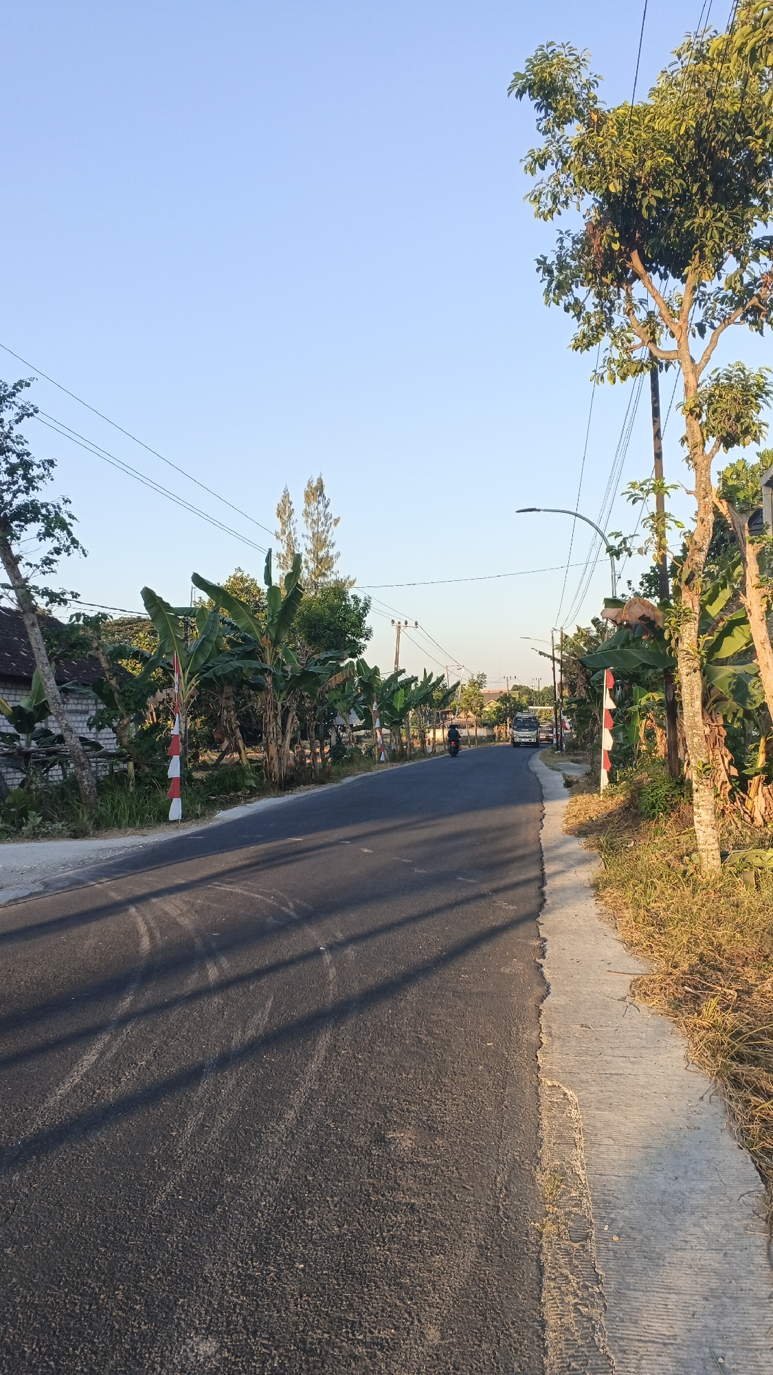 The image depicts a rural road in Soko, Indonesia, bathed in the warm light of a clear morning. The asphalt road curves gently, flanked by a concrete sidewalk on the right and a grassy verge. To the left, banana trees and other vegetation line the roadside, with a glimpse of a brick wall and roof of a building. Red and white striped markers are visible along the edge of the road, likely delineating boundaries or serving as visual aids. Power lines crisscross the sky, supported by utility poles. A lone motorcycle rider is seen on the road in the distance, and a white vehicle approaches from further down the road. The sky is a soft, pale blue, indicating early morning or late afternoon.