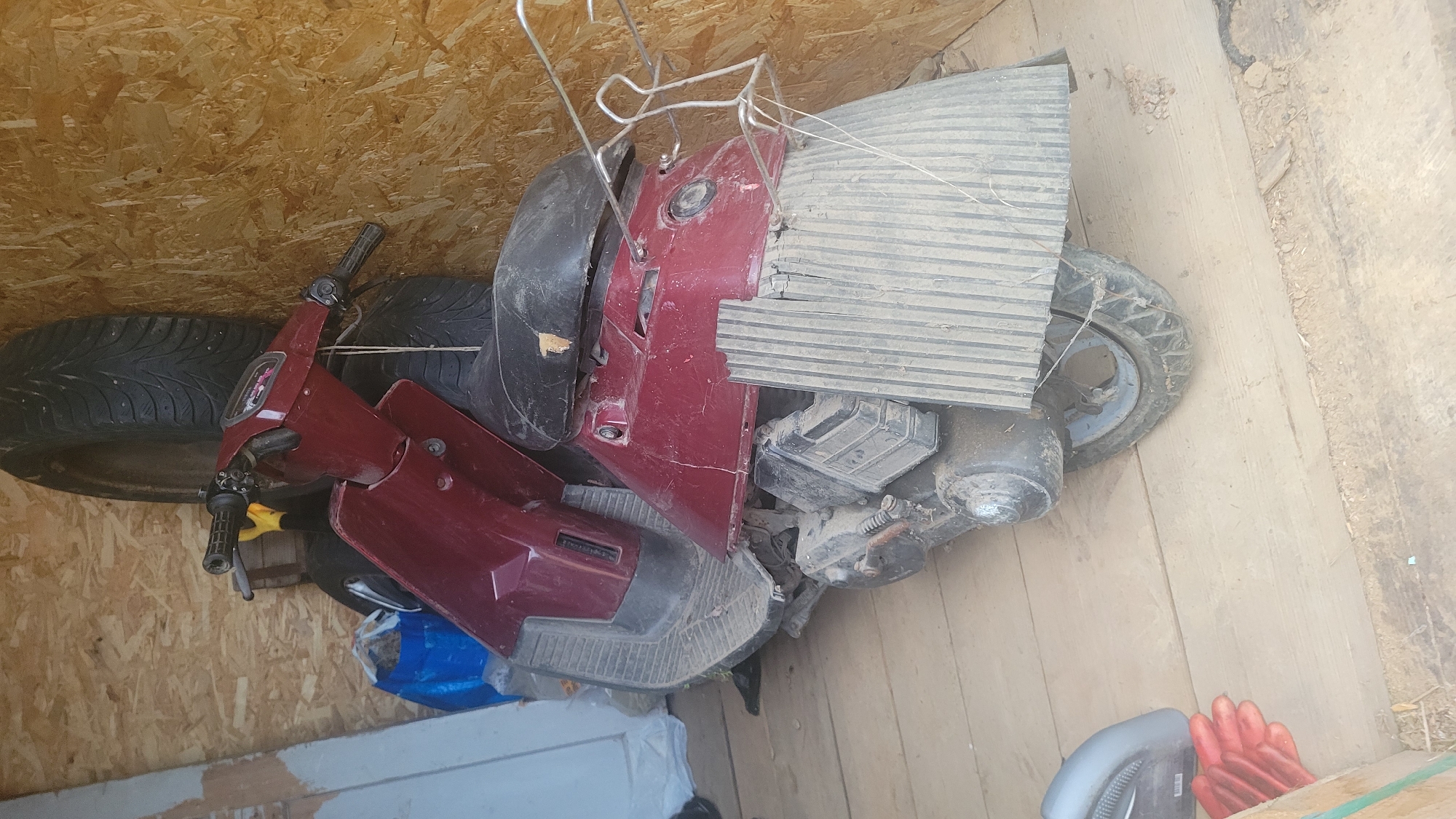 A red scooter is parked on a wooden floor inside a storage unit. The scooter is covered in dust and cobwebs. Behind the scooter, two large tires are leaning against a wall made of particleboard. To the left of the scooter, a blue plastic bag is visible. In the foreground, a pair of red gloves lies on the floor. The overall environment appears to be a cluttered storage space.