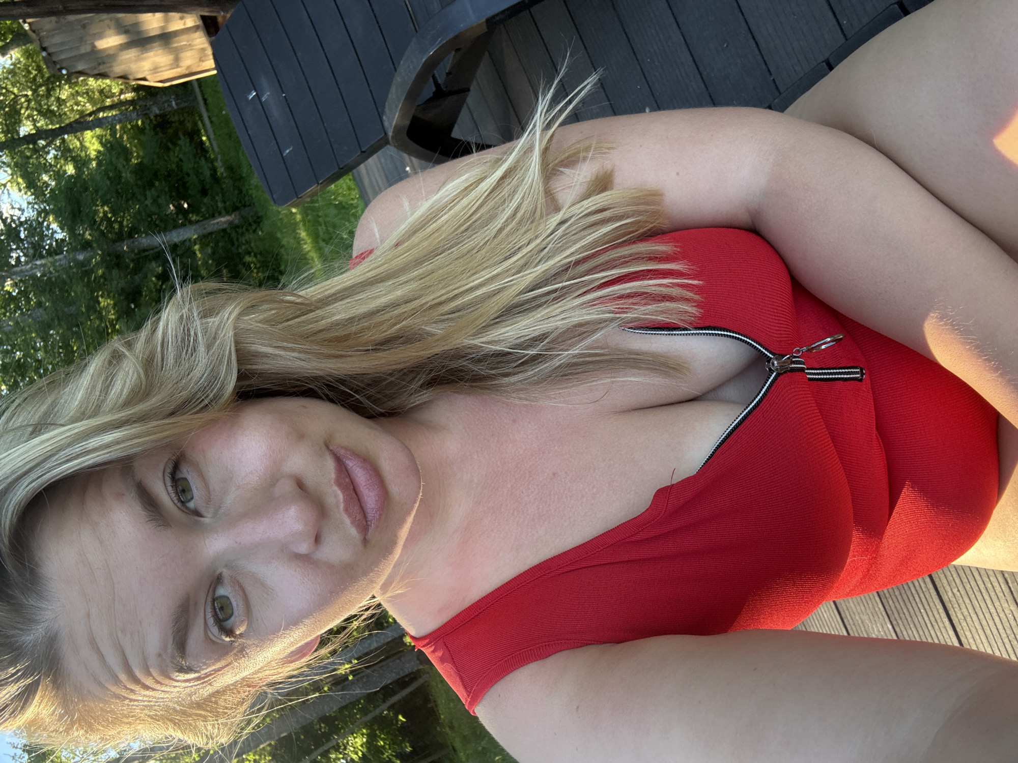 A blonde woman is taking a selfie outdoors. She is wearing a red zippered swimsuit. She is seated on a wooden deck. Behind her are trees and a wooden structure. The scene appears to be in a rural or natural setting, potentially in or around Zamosh'e (Zaklinskoe S/P), Russia, given the presence of trees and a wooden cabin-like structure. The lighting suggests it is daytime, possibly late afternoon or early evening due to the warm, golden light.