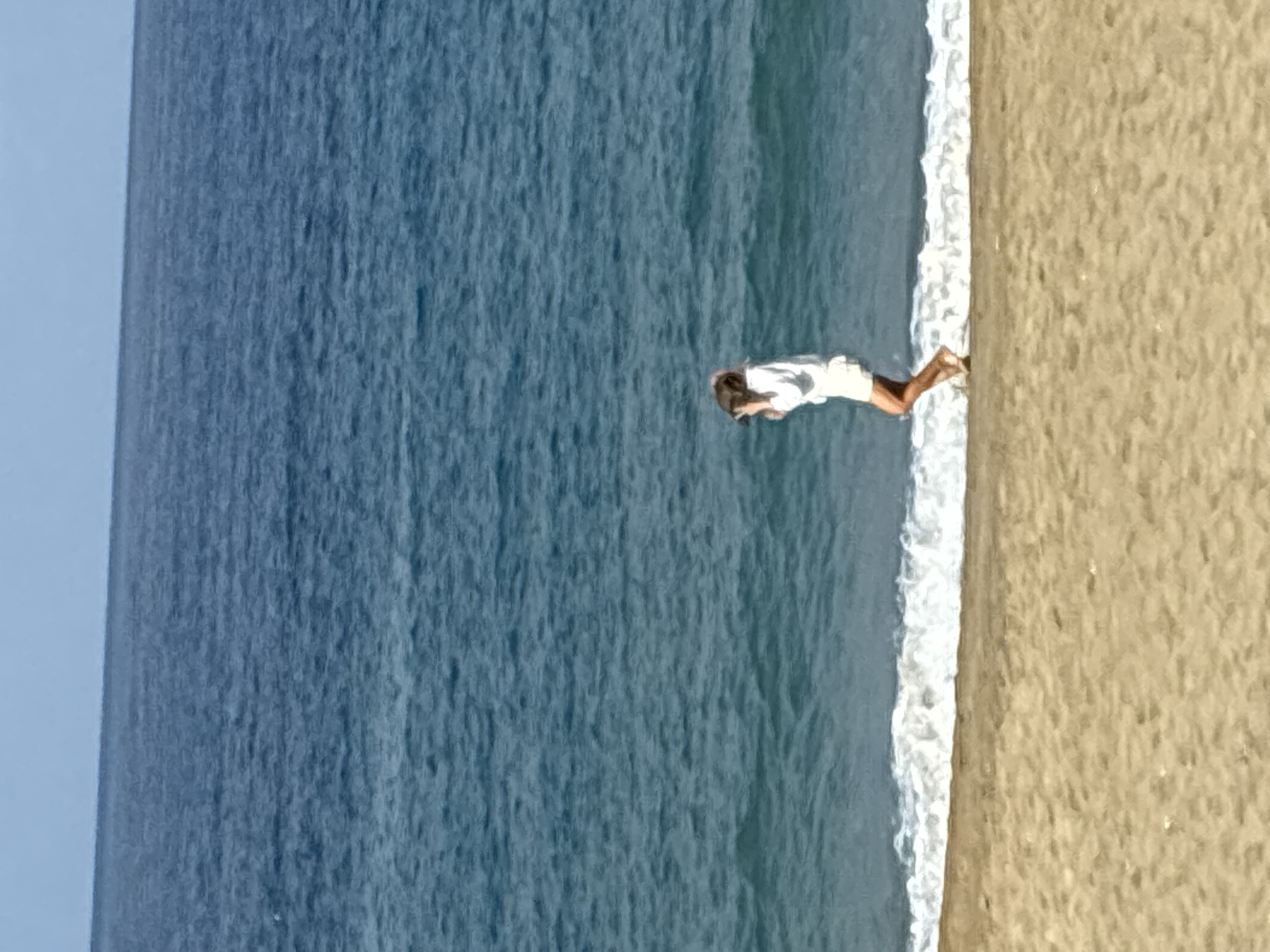 An individual is captured mid-stride on a sandy beach, with the turquoise waters of the Mediterranean Sea stretching out behind them, meeting the horizon under a pale blue sky. The person, wearing a white top and shorts, appears to be holding a camera to their face, possibly in the act of taking a photograph or recording a video. The waves are gently breaking on the shore, creating a white foamy line where the water meets the sand. The sand itself is a light brown, with some small debris visible. The scene is one of leisure and activity by the sea. The location is likely a public beach in Alanya, Turkey, a popular coastal city known for its Mediterranean shoreline.