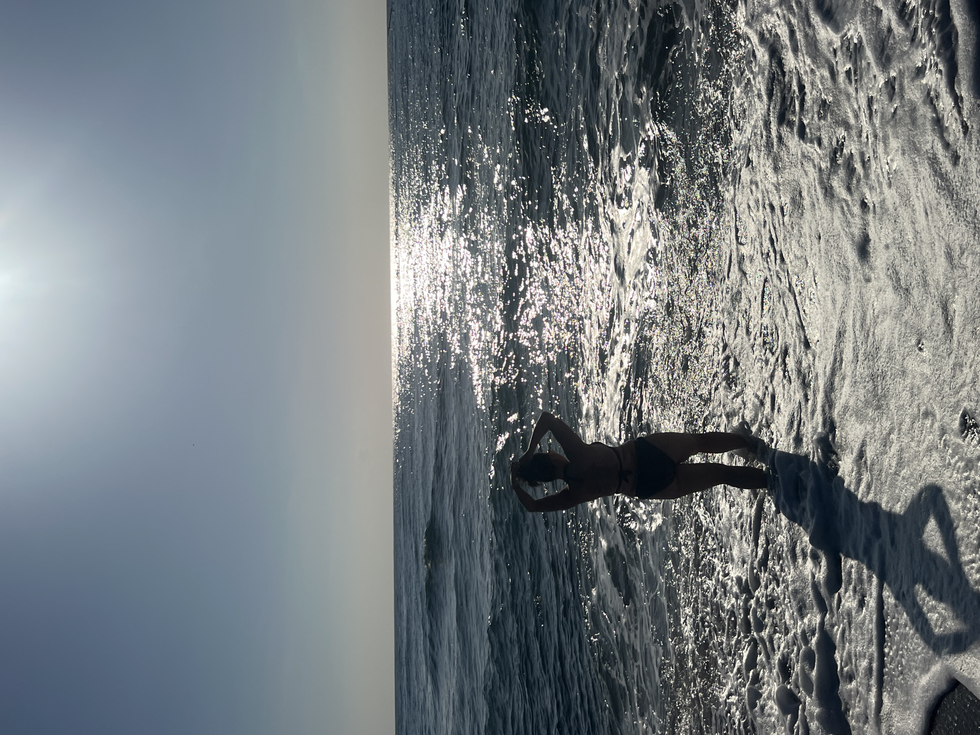 The image shows a person from behind, standing at the edge of the ocean, with waves washing over their feet. The person is wearing a dark bikini and has their hands up, touching their hair. The sun is bright and reflecting off the water, creating a shimmering effect. The sky is a clear blue, with a slight haze near the horizon. The overall scene is one of a serene beach setting. The location is not specified as Abang, Indonesia.