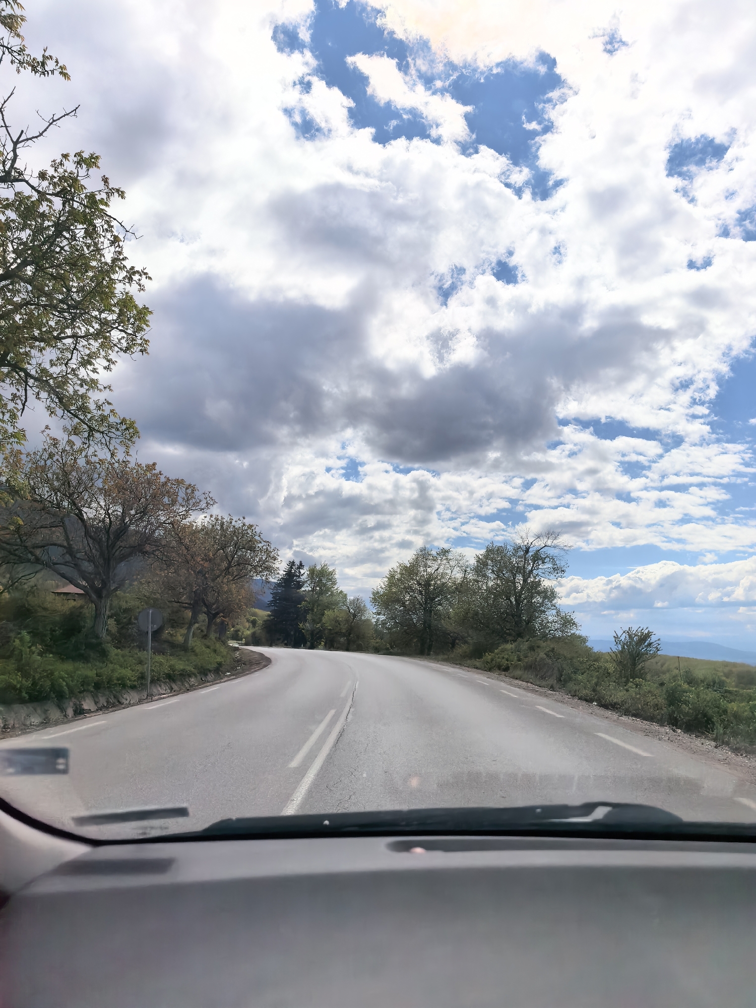 A view from inside a vehicle looking out onto a paved road that curves slightly to the left. The road has painted white lines along each side and a broken white line down the middle. Trees and shrubbery line both sides of the road, with some trees still bare and others beginning to leaf out. In the distance, a hint of a landscape is visible. The sky is filled with a mix of white and gray clouds with patches of blue. The dashboard of the vehicle is visible at the bottom of the frame, partially obscuring the view of the road immediately in front of the car.
The location of this image is on the way to the Seven Rila Lakes from Sapareva Banya, Bulgaria.