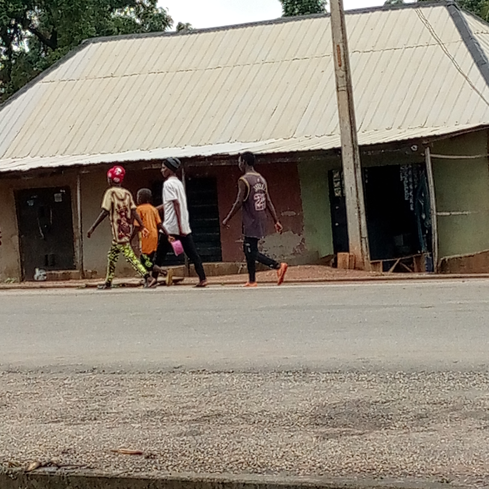 In Mararaba, Nigeria, four people are walking on a road in front of a building. The building is one-story, with a corrugated metal roof, and has several storefronts with closed doors. An electrical pole stands in front of one storefront. The road is paved. Of the four people, one is wearing a red helmet and printed shirt with lime green leggings, another is wearing an orange shirt, the third is wearing a white shirt, and the fourth is wearing a basketball jersey with black pants.