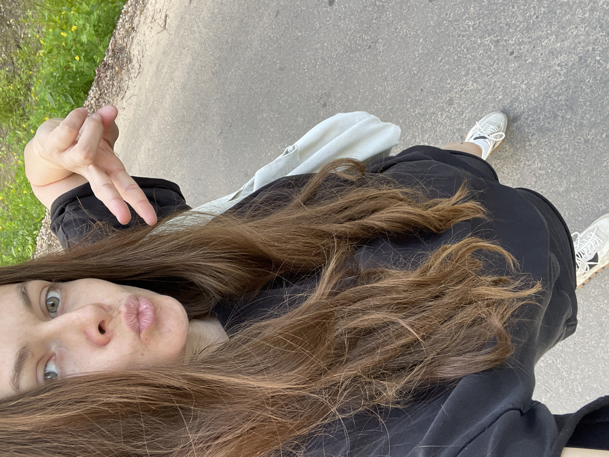 The image shows a selfie taken from a high angle, looking down at the subject and the surrounding environment. The subject is a young woman with long, brown hair that cascades over her shoulders and partially obscures her face. She has light green eyes and is wearing a black outfit. Her lips are pursed in a playful expression. She is holding up her left hand with two fingers extended, forming a peace sign.
In the background, a paved path is visible, suggesting an outdoor setting. To the left of the path, there is a patch of green grass with some yellow flowers, indicating a natural environment, possibly a park or a roadside. A white bag is placed on the ground next to the subject. Her white sneakers are also visible, suggesting she is standing or walking on the path.
The lighting suggests it is daytime, with soft natural light. The overall mood of the image is casual and cheerful. The location is identified as Zarechny, Russia. There is no visible text in the image.
