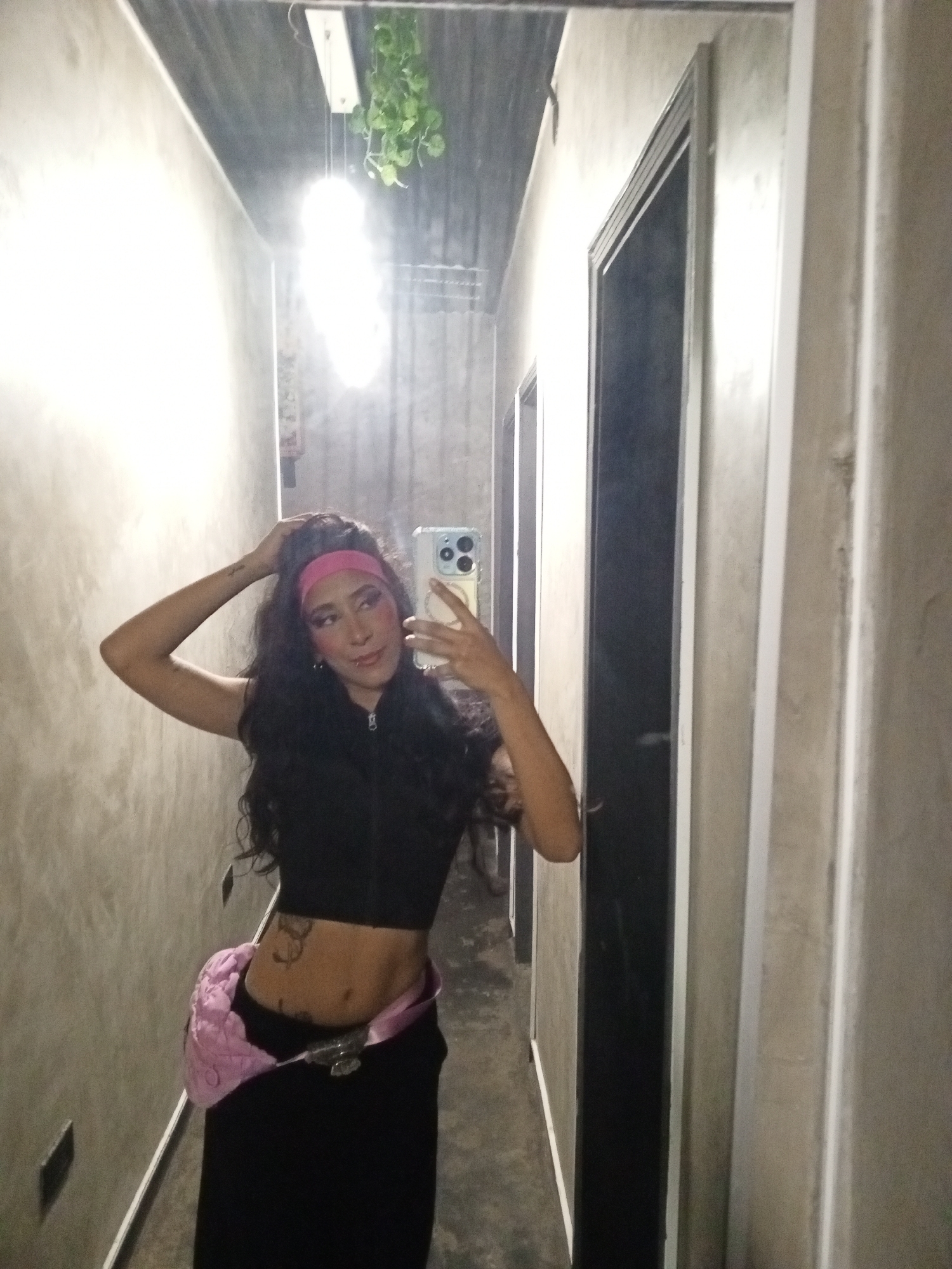A woman is standing in a narrow hallway, taking a selfie in a mirror. She has dark, curly hair and is wearing a pink headband, a black crop top, and a black skirt. She has a pink fanny pack around her waist. The hallway has grey walls and a grey ceiling. There is a light fixture with a hanging plant on the ceiling. There is a door in the hallway on the right. The location appears to be an interior space, possibly a residential building.