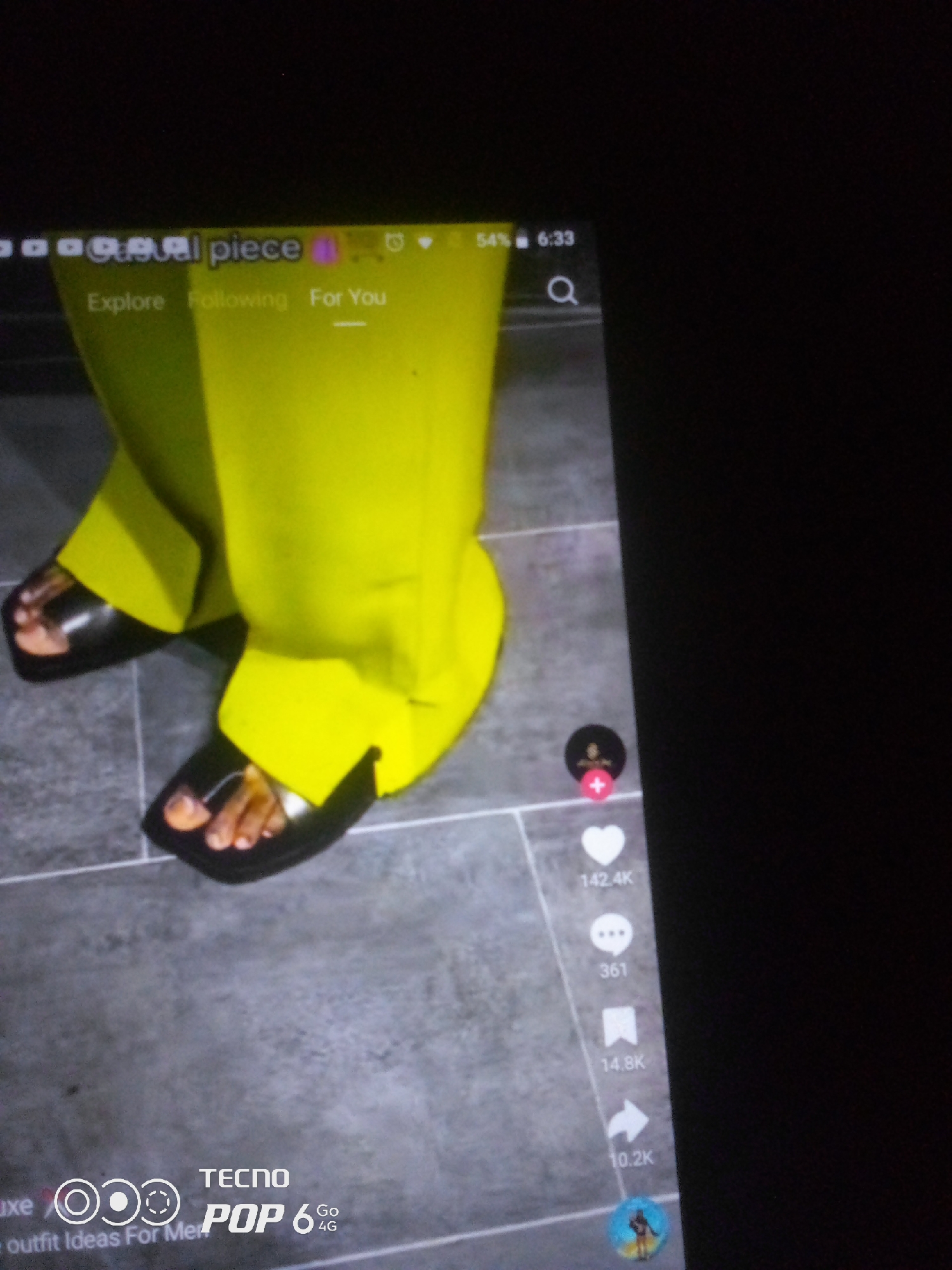 The image displays a close-up view of a person's feet wearing black square-toed slides and bright yellow wide-leg pants. The background appears to be a tiled floor. The scene is captured on a screen, likely a smartphone, as evidenced by the status bar at the top showing "54% 6:33" and icons for explore, following, and for you. There are also social media engagement icons on the right side of the screen, including a heart with "142.4K" likes, a speech bubble with "361" comments, a bookmark with "14.8K" shares, and an arrow with "10.2K". In the lower left corner, text reads "luxe" and "outfit Ideas For Me," alongside a TECNO POP 6 4G logo. The image is not located in Howayi, Nigeria, as there is no information in the visual or text to support this claim.