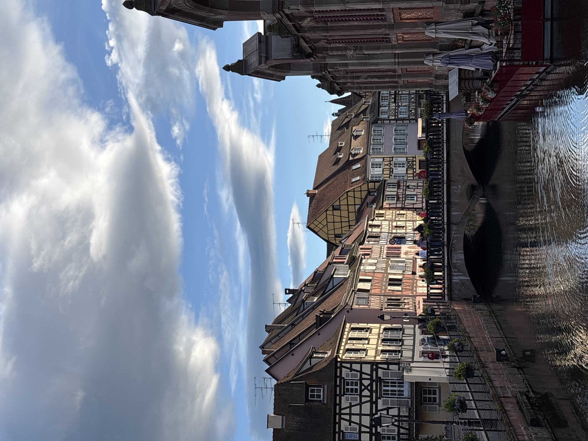 A vertical, eye-level medium shot captures a picturesque canal scene in Colmar, France, under a dramatic sky filled with textured clouds and patches of blue. The canal, reflecting the light, flows from the foreground toward the background, flanked by quaint, colorful half-timbered buildings. On the left bank, a stone wall lined with lampposts and hanging flower baskets separates the canal from a walkway. Several people are visible on this walkway, some walking, others standing and conversing. A brick bridge with an arched underside spans the canal, its top serving as a pedestrian walkway. More people are gathered on the bridge, observing the scenery. On the right bank, a building with large arched windows and intricate stonework dominates the right side of the frame. In front of this building, a red-railed outdoor seating area with tables, chairs, and closed umbrellas suggests a cafe or restaurant. The architecture of the buildings is characteristic of Alsace, with steeply pitched roofs and decorative half-timbering. The overall impression is one of a charming, historic European town.