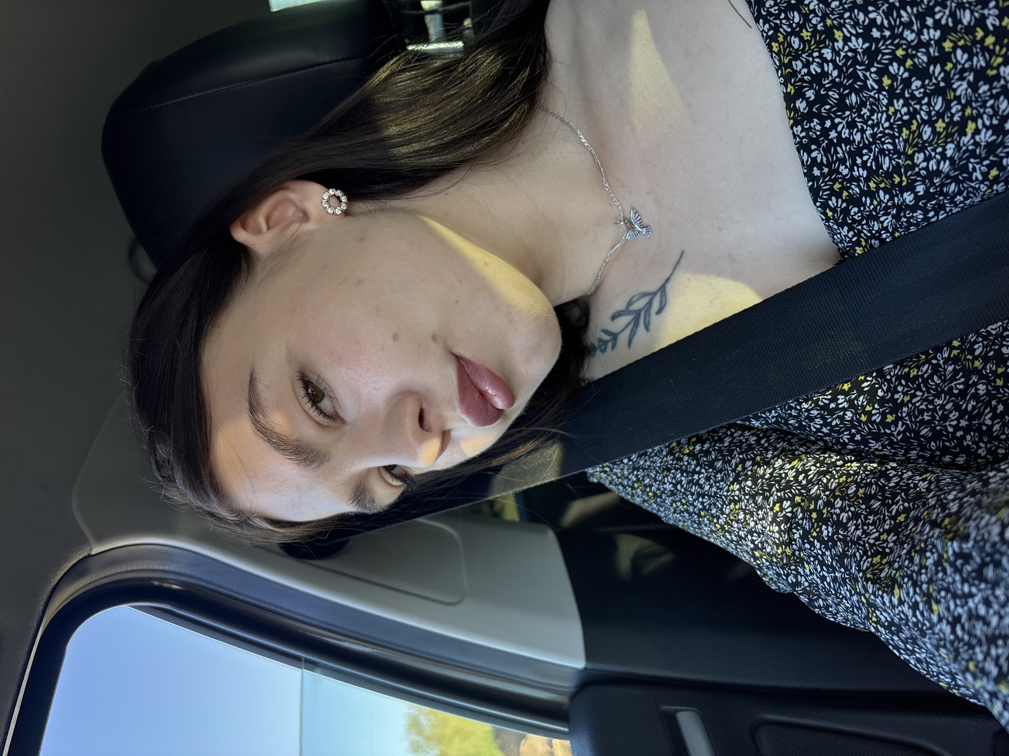 A woman is in the passenger seat of a car. She is wearing a floral print dress, a silver necklace with a bird pendant, and a seatbelt. She has an earring in her left ear, a tattoo on her chest, and dark hair. The car is moving; the view outside the window is blurred. The location is Perm, Russia.