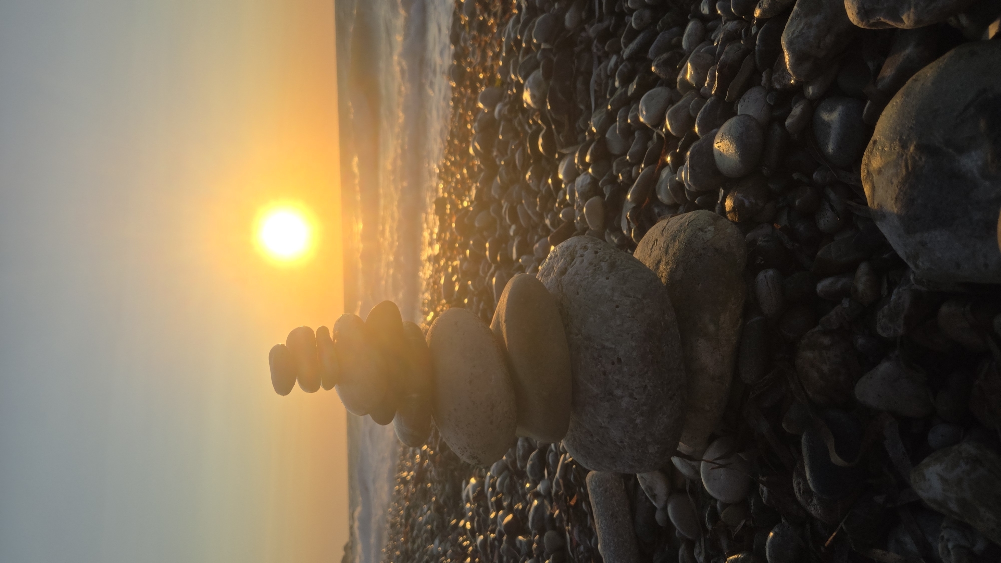 A stack of smooth, grey stones is balanced on a rocky beach at sunset in Soroni, Greece. The sun, a bright, luminous orb, is positioned in the upper right quadrant of the image, casting a warm, golden glow across the scene. The waves of the Aegean Sea gently lap at the shore in the background, their crests catching the sunlight. The beach itself is covered in a dense layer of pebbles and stones, some of which are wet and glistening from the receding tide. The sky is a gradient of soft oranges and yellows, transitioning to a pale blue at the top. The overall atmosphere is serene and tranquil.