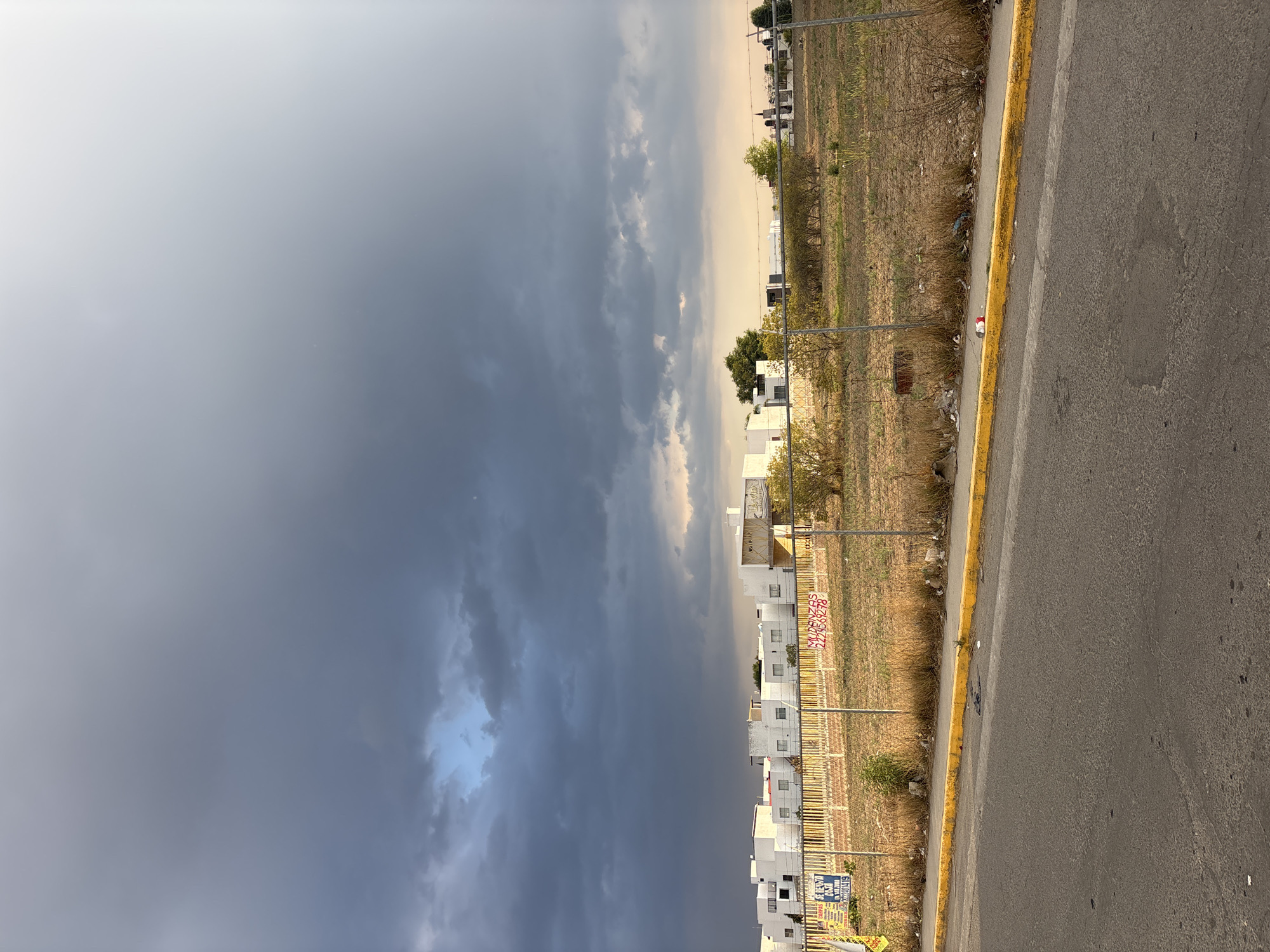 The image depicts a view along a road in San Juan Cuautlancingo, Mexico. A line of white buildings, likely residential, stands behind a metal fence. Between the fence and the road is a strip of dry, grassy land. The road itself is asphalt, marked with a yellow line. The sky is overcast, with dark storm clouds dominating the view.