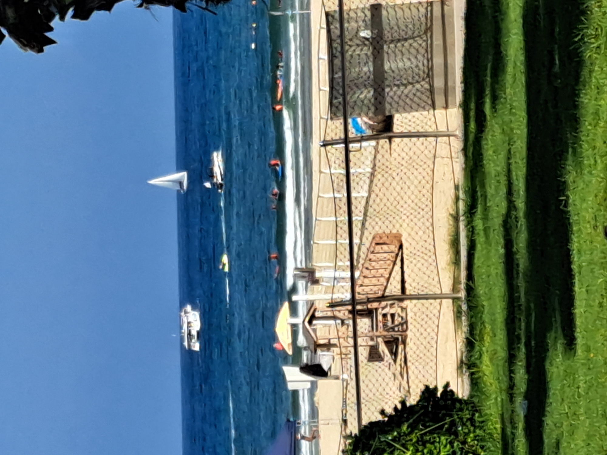 The image depicts a sunny day at a beach in Herzliya, Israel. The sea is a vibrant blue with moderate waves, and several boats are visible on the water. A white sailboat with a single sail is further out, while a white speedboat with people on board is closer to shore. A larger white boat, possibly a ferry or tour boat, is also in the distance. Closer to the shore, there are people engaged in water activities, including some on inflatable rafts or bodyboards.
The beach itself is sandy, with a playground structure featuring a slide and wooden steps. A yellow umbrella provides shade near the playground. A portion of a blue tent is visible on the far left. A chain-link fence separates the beach area from a grassy foreground, where shadows are cast by unseen trees or structures. The overall scene conveys a lively atmosphere of a popular beach resort.