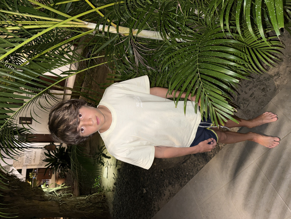 A young person stands amongst palm fronds and greenery. They are wearing a light-colored t-shirt and dark shorts with a yellow stripe, standing barefoot on a paved surface. Building details are visible in the background, including lit windows and signage, possibly of a hotel. The scene is illuminated at night.