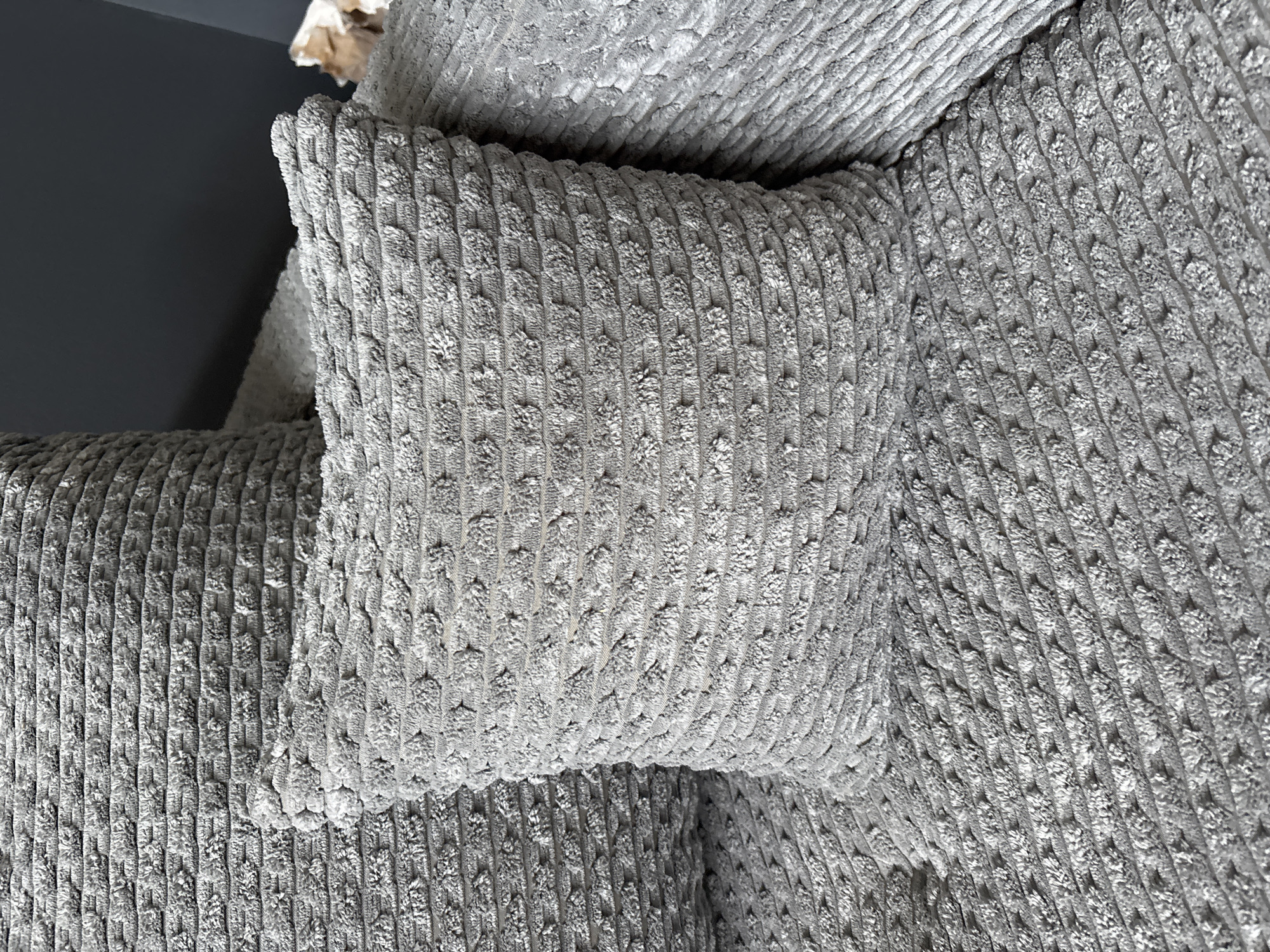 The image shows a close-up view of a grey corduroy fabric, likely a sofa or a cushion. The texture of the fabric is characterized by raised parallel ribs and a nubby, textured surface within each rib. The lighting creates shadows that highlight the three-dimensional nature of the weave.
The scene is interior, with a dark grey wall visible in the upper left corner, suggesting a room setting. The fabric is draped and folded, creating soft curves and revealing different angles of its textured surface. There are no people or explicit activities depicted. The overall impression is one of comfort and softness, due to the plush appearance of the fabric.
The location context of Zaporizhzhia, Ukraine, does not provide any specific visual cues within the image itself. The time of day and weather are also not discernible from the image. There is no visible text. The focus is solely on the material and its texture.