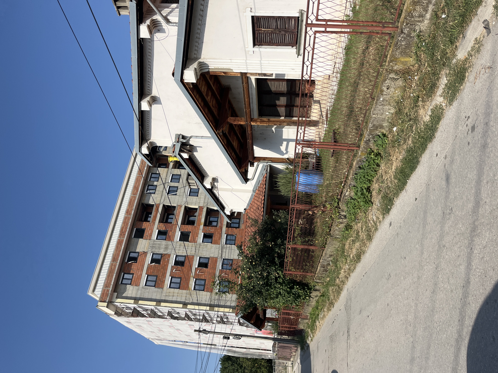 The image shows a street scene in the city of Cacak, Serbia. In the foreground, a paved road runs diagonally from the bottom left towards the center right of the frame. The road appears to be in fair condition, with some wear and tear visible. To the right of the road, a concrete embankment lines the street, topped by a rusty metal fence. Beyond the fence, a white house with a wooden-roofed porch is visible. The house has dark-shuttered windows and appears to be older. Behind the house and extending to the left of the frame is a large, multi-story building under construction. This building is partially covered in white sheeting and has exposed brickwork and concrete. Several windows are visible, some of which are open. A cluster of trees and greenery can be seen behind the fence and in the distance. A bright blue barrel is placed near a small covered structure with a red tiled roof, situated between the house and the construction site. The sky is clear and blue, indicating a sunny day. There are no people visible in the image, and the scene depicts a typical urban or suburban environment undergoing development.
