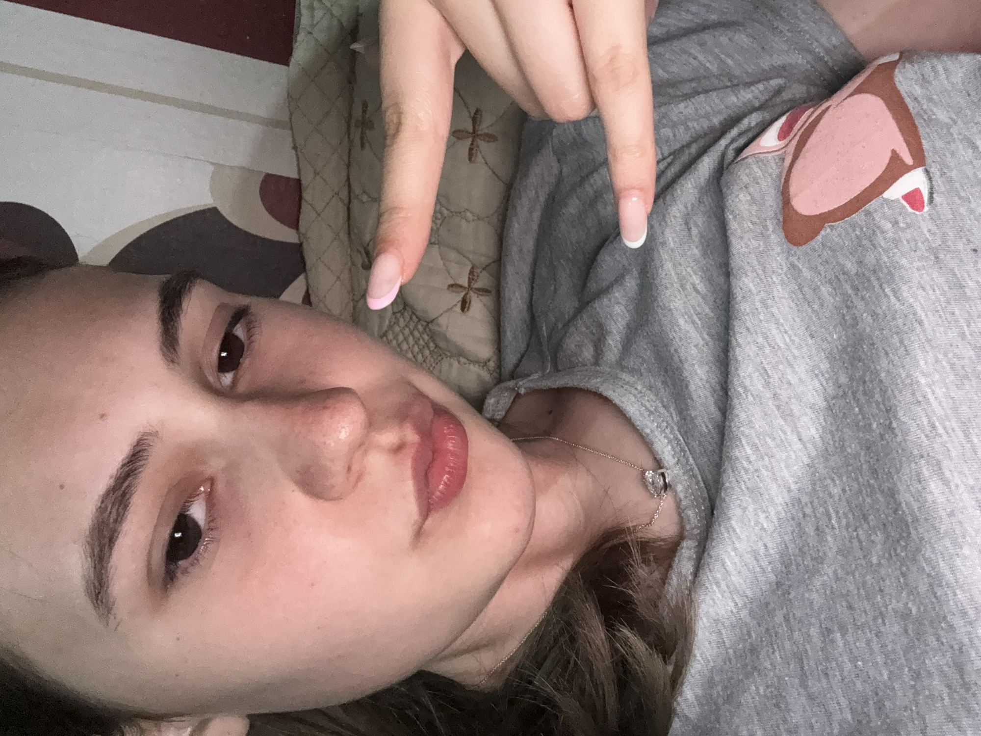 A young woman is lying down, possibly on a bed, judging by the patterned bedding and pillow visible. She is fair-skinned with brown hair and eyes. She is wearing a grey t-shirt with a brown and pink animal ear graphic on the shoulder. She is also wearing a delicate silver necklace with a heart-shaped pendant. Her nails are manicured with a French tip, with one hand displaying a pink tip. There is no clear information that indicates the location is Panagyurishte, Bulgaria.
