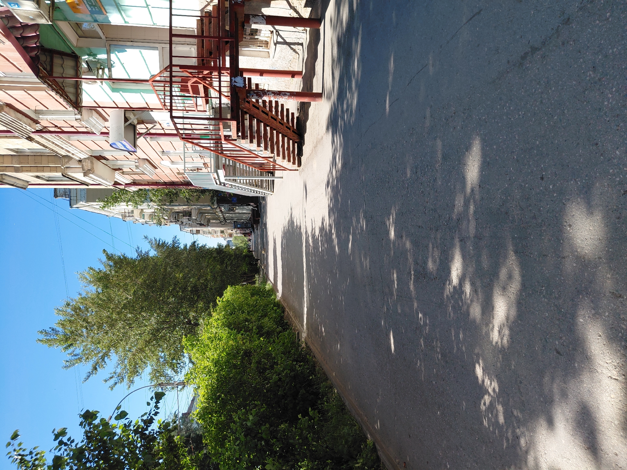 A street view in an urban setting shows a paved sidewalk adjacent to a row of buildings and a dense green hedge. The buildings are multi-story structures with facades featuring windows and balconies. Red metal external staircases are visible attached to some of the buildings. Trees line the street, casting dappled shadows on the pavement. The sky above is clear and blue.
