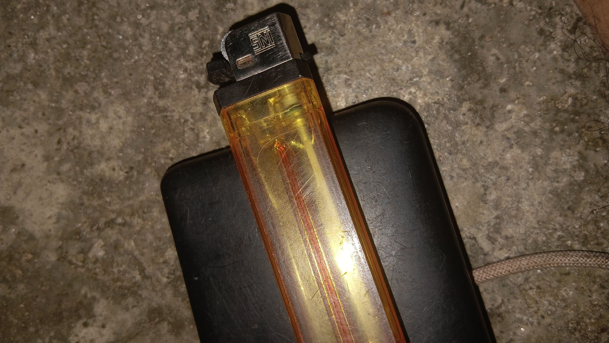 A disposable butane lighter with a translucent orange body and a silver metal top is placed on a black, rectangular object, possibly a power bank. The lighter has a silver wheel mechanism for ignition and a visible fuel chamber with red-colored elements. The objects are resting on a rough, gray, textured surface, likely concrete, with some scattered debris. The background is dark and out of focus, with a small section of coarse, light-brown woven cord visible on the right. The scene is lit from above, casting subtle shadows. This scene is not specific to any location in the city of Prabumulih, Indonesia.