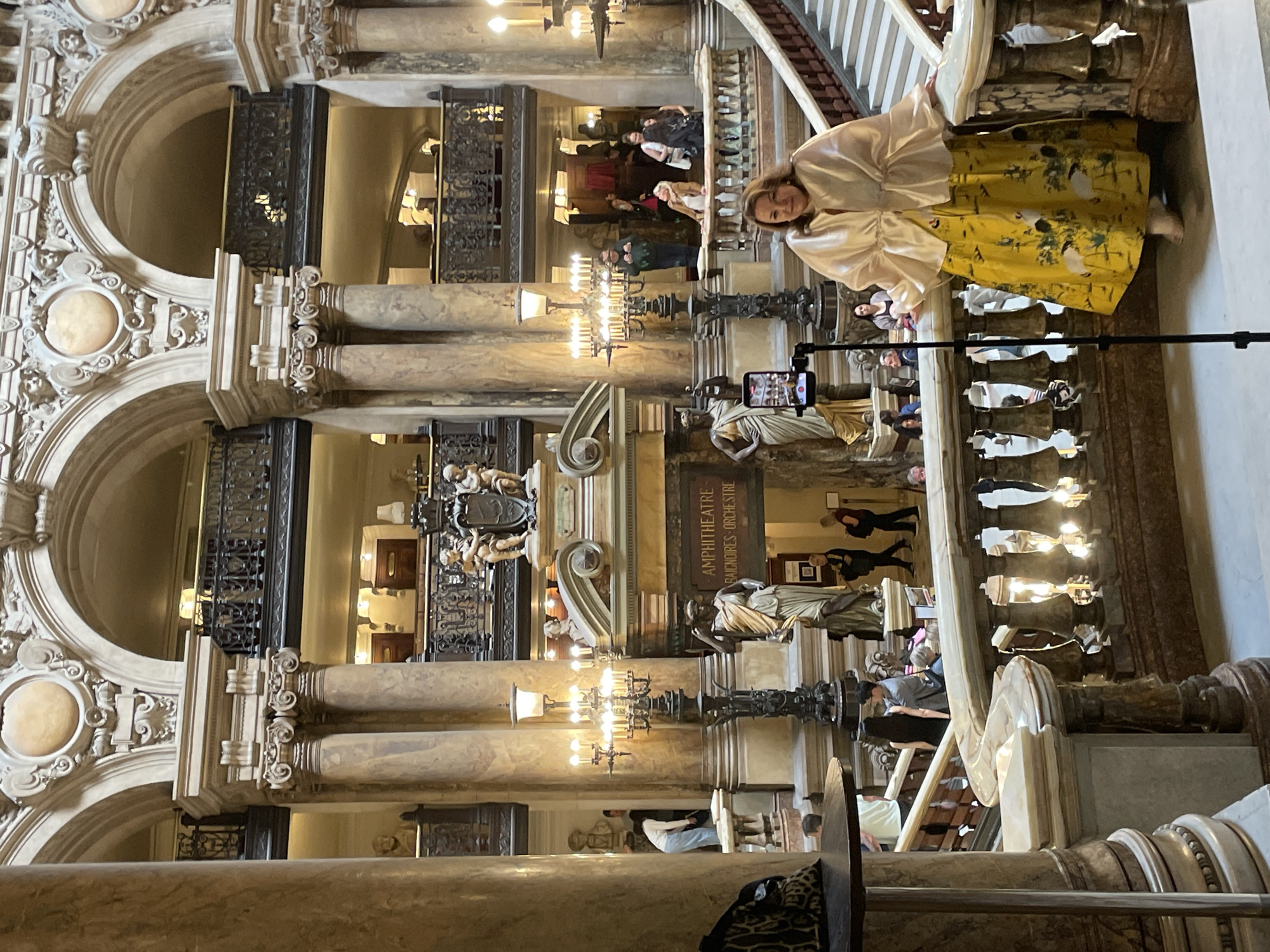This media file captures an interior view of a grand, ornate building, likely a theatre or opera house, given the architectural style and signage. The scene is brightly lit, suggesting it's daytime. A woman in a light-colored, billowy top and a yellow skirt adorned with a bird and floral pattern stands on a balcony, posing for a photo taken by a smartphone mounted on a tripod. She appears relaxed and engaged with the photography setup.
Below the balcony, other individuals are visible, some walking or standing, suggesting the space is open to visitors or patrons. The architecture is characterized by marble columns, intricate carvings, and wrought-iron railings, indicative of a historic and opulent European setting, possibly Paris, France. Chandeliers and other decorative lighting fixtures are also present.
A prominent sign reads "AMPHITHÉÂTRE BAIGNOIRES-ORCHESTRE," further confirming the location as a performance venue and providing specific section names. The overall atmosphere seems to be one of leisurely exploration and appreciation of the building's grandeur. The detailed carvings and the formal layout suggest a rich history and cultural significance to the location.
