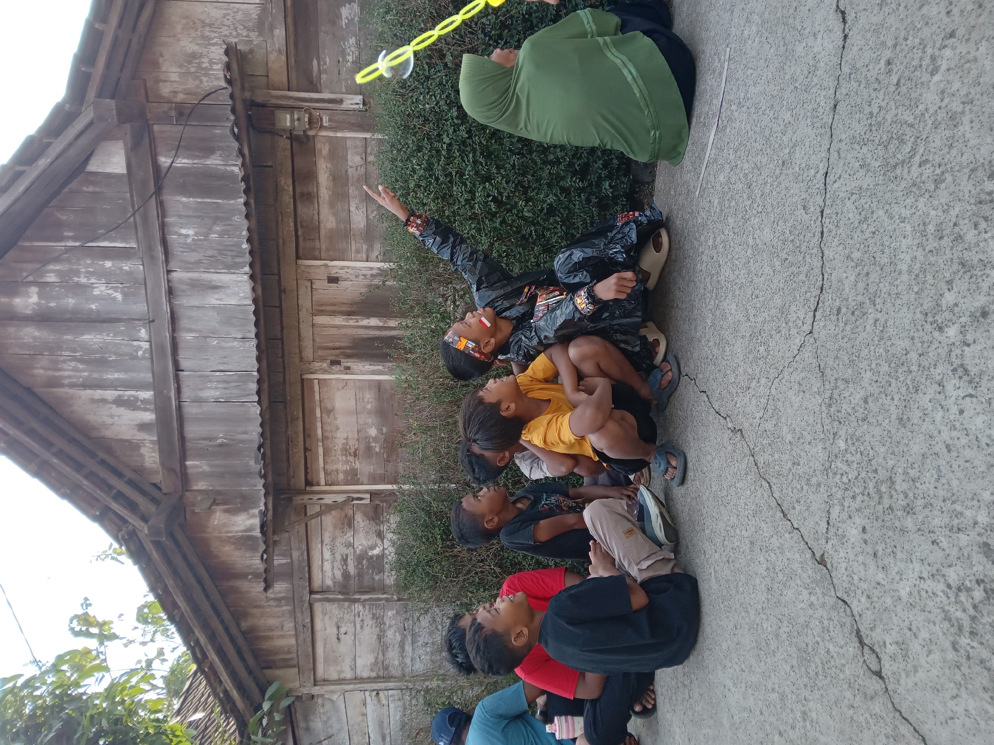 In Kedunggudel, Indonesia, a group of children and an adult sit on a concrete surface in front of a weathered wooden building. The building has a corrugated metal roof and appears to be old. A hedge runs along the base of the building. Some of the children are looking up at something, while one boy, dressed in a black plastic-like costume with colorful trim, points upwards. A person wearing a green hijab is also present, seated to the right of the boy in the costume. A yellow bubble wand with bubbles is visible in the upper right portion of the image, suggesting the children are engaged in blowing bubbles. The ground is cracked concrete, and some foliage is visible in the background. The scene appears to be an outdoor gathering in a village or rural setting.