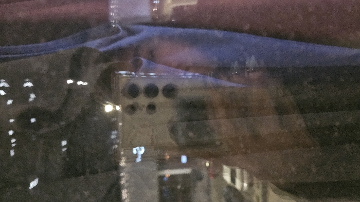 The image shows a blurry reflection inside a car window, likely at night. Visible through the reflections are city lights and buildings. A device with several circular openings, possibly a camera or electronic gadget, is also reflected in the window, along with what might be the driver's head silhouette. The window itself has horizontal defroster lines visible.