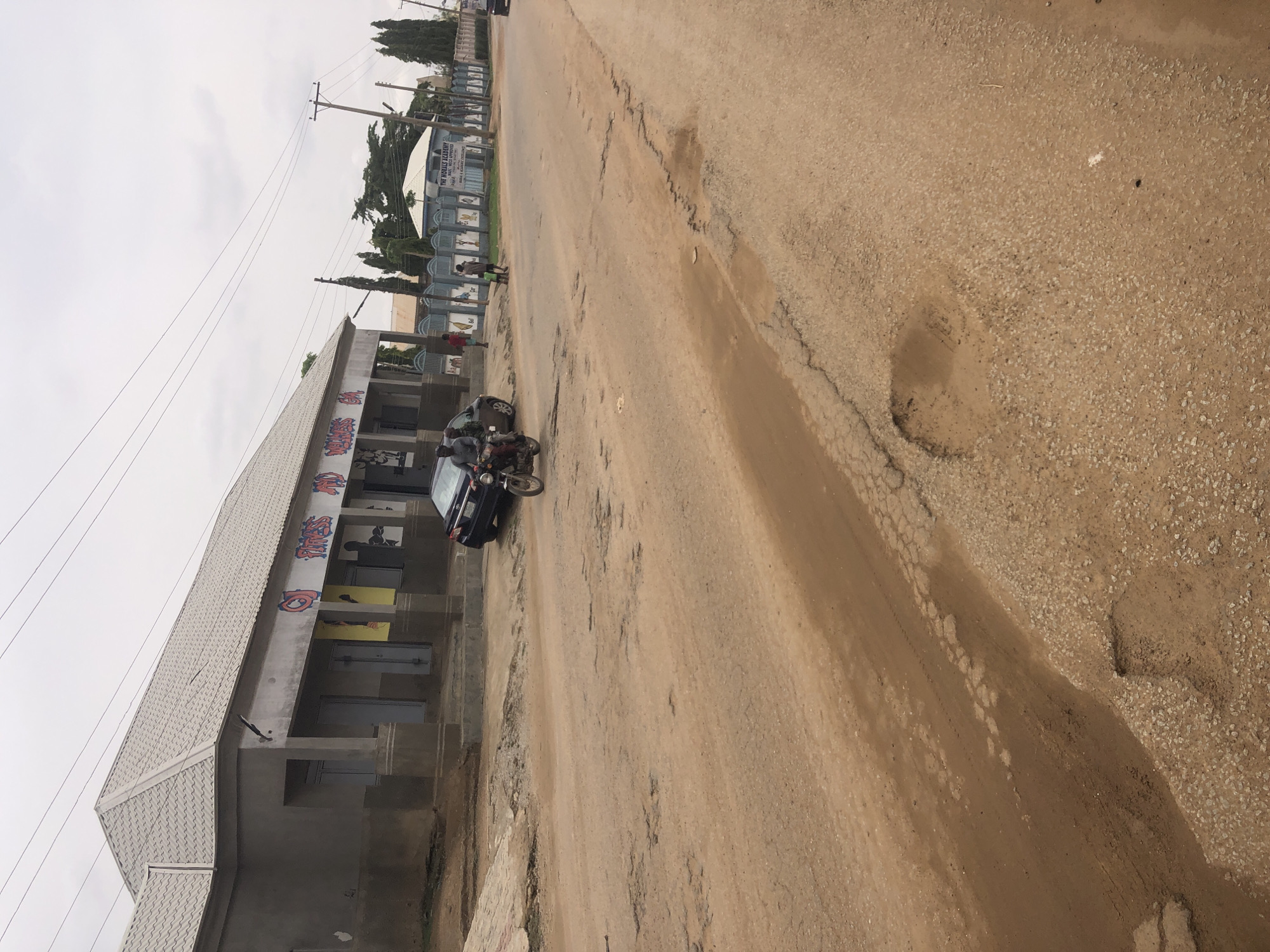 The image shows a street scene in Hanwa, Nigeria. The road is unpaved, with significant damage and potholes. A building with a light-colored roof and painted murals is visible along the side of the road. A dark-colored car is parked near the building, and a motorcycle with two riders is positioned next to it. Several people are walking along the sidewalk in the distance. Overhead electrical wires are visible. The sky is overcast.