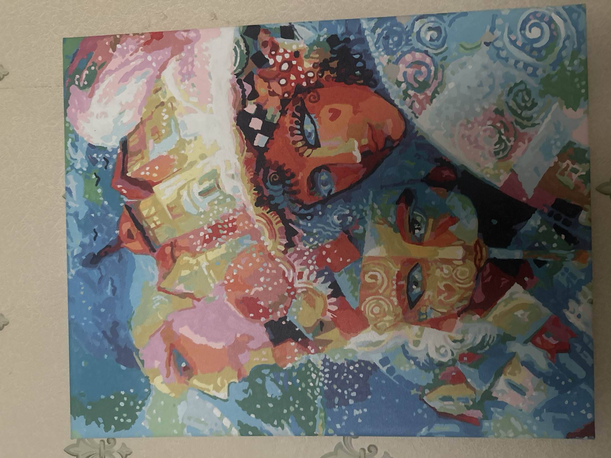 This is a photograph of a painting. The painting is an abstract depiction of what appear to be three faces, possibly women, rendered in a vibrant and expressive style. The faces are intertwined and overlap, creating a sense of depth and movement. The color palette is rich and diverse, featuring blues, oranges, reds, yellows, pinks, and white. There are various patterns and textures within the painting, including dots, swirls, and checkered elements, adding to its abstract and artistic nature. The faces have distinct features like eyes and noses, but they are stylized and not anatomically precise. The background is also abstract, with a mix of swirling patterns and solid colors. The overall impression is one of energy and emotion, possibly representing a dreamlike or surreal scene. The painting is displayed on a wall with a textured wallpaper. There is no visible text in the image.