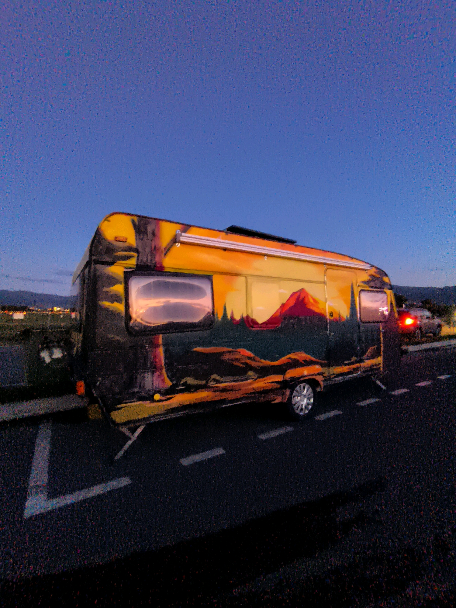 The image shows a camper van parked on the side of a road. The camper van is decorated with a mural depicting a mountain landscape with a sunset. The sky is a deep blue, indicating it is either dusk or dawn. There are road markings on the asphalt. In the background, there are lights from buildings and a car with its brake lights on. The scene appears to be on the outskirts of a city, with some agricultural fields visible on the left. The overall lighting is dim, suggesting low light conditions. The image does not provide any specific visual cues to confirm the location as Batumi, Georgia.