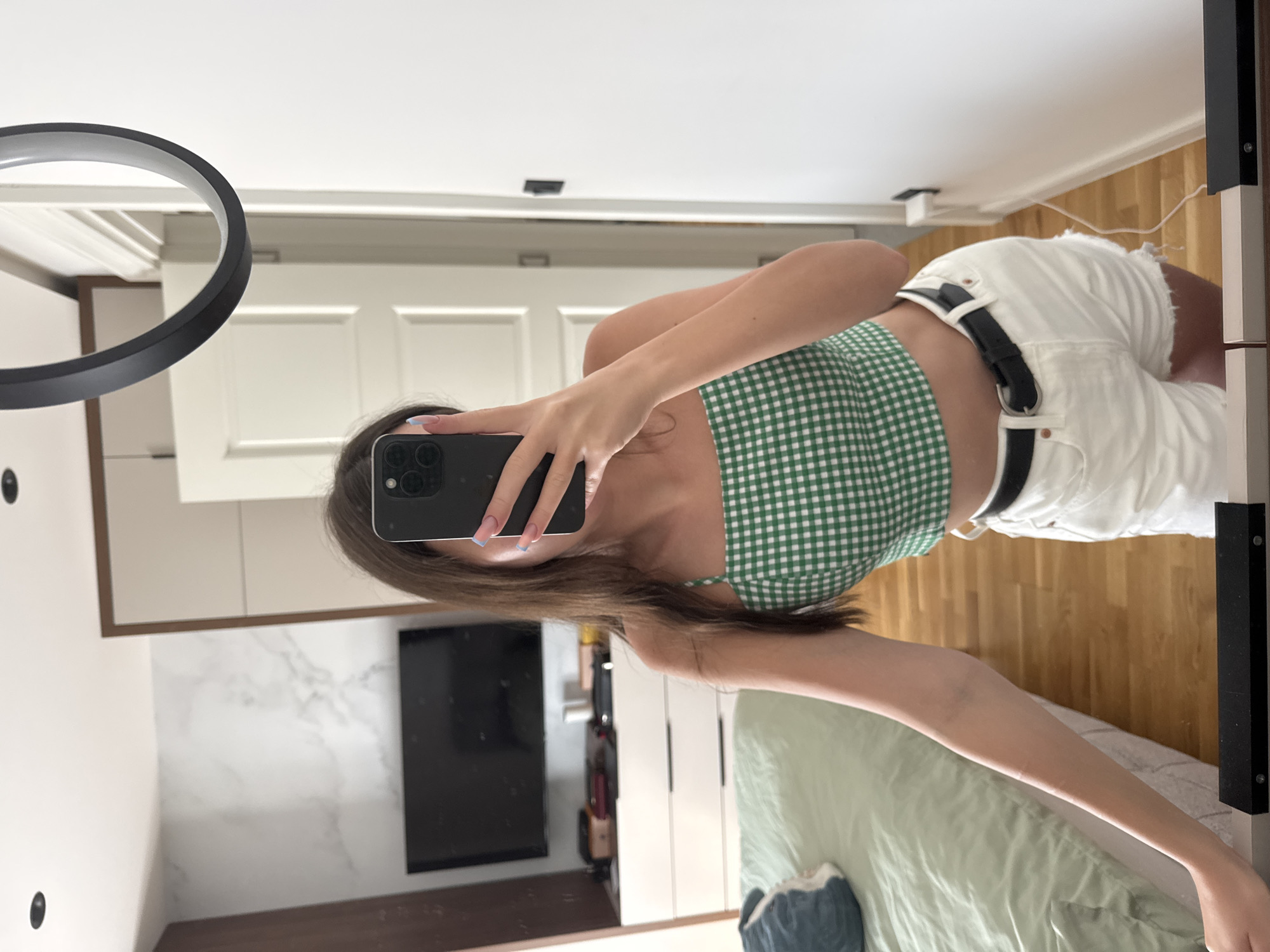 A young woman is reflected in a mirror, taking a selfie. She is wearing a green and white checkered crop top and white denim shorts with a black belt. Her long brown hair is down and covers part of her face. In the background, a bed with a light green comforter is visible, along with a dresser and a television mounted on the wall. The room appears to be a bedroom.