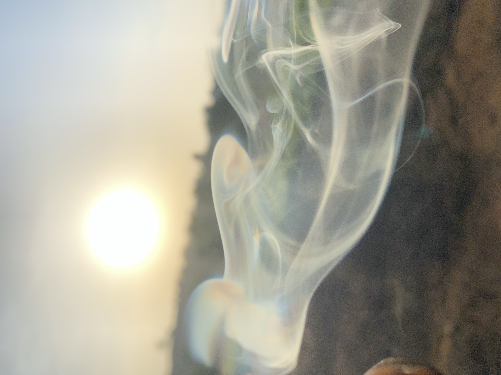 The image presents a blurred scene with a prominent wisp of smoke centrally positioned. To the left, a bright, out-of-focus circular light source is visible, possibly the sun. The background is a mix of blurred light and dark areas. To the right of the smoke, a textured, dark surface is present. The location of Amancho, Nigeria, is not discernible from the image alone.