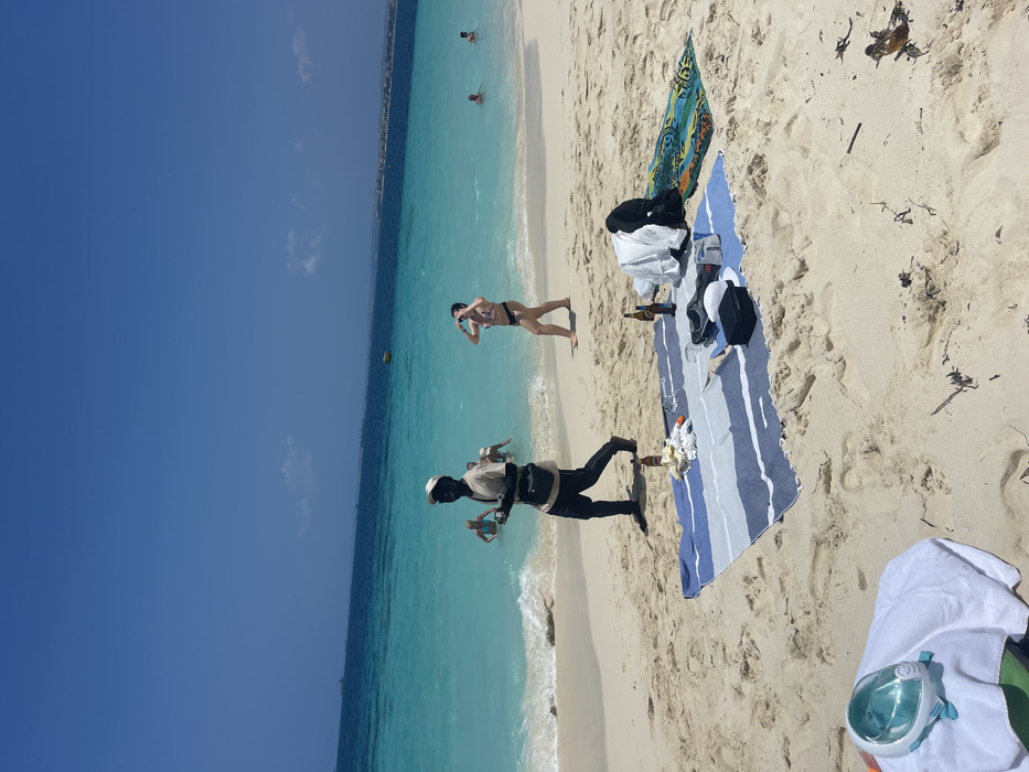 The image depicts a sunlit beach scene in Zanzibar, Tanzania. The sky is clear and blue with sparse white clouds. The ocean water transitions from light turquoise near the shore to deeper blue further out, with small waves breaking on the sandy coast.
In the mid-ground, a dark-skinned male, wearing a light-colored long-sleeved shirt, dark trousers, dark shoes, and a dark baseball cap, walks from left to right across the sand. He carries several small, light-colored figures in his left hand, holding them up, and a dark cross-body bag hangs from his shoulder. He looks directly forward, towards the viewer. To his right, a light-skinned female in a dark two-piece bikini walks out of the shallow water onto the sand, her arms slightly raised. Further in the water to the right, three other individuals are visible swimming or wading. A yellow buoy floats in the mid-distance water.
On the foreground-midground sand, a blue and white striped beach towel is spread. It holds two brown glass bottles, a crumpled white item, a white garment, a pair of dark shoes, and a small black rectangular container. Adjacent to this towel, a colorful patterned beach mat is laid out. In the lower-left foreground, a white and light blue full-face snorkeling mask rests on a white towel. The light-colored sand exhibits footprints and scattered dark organic debris. On the distant horizon, a landmass with several visible structures and multiple vessels are present.