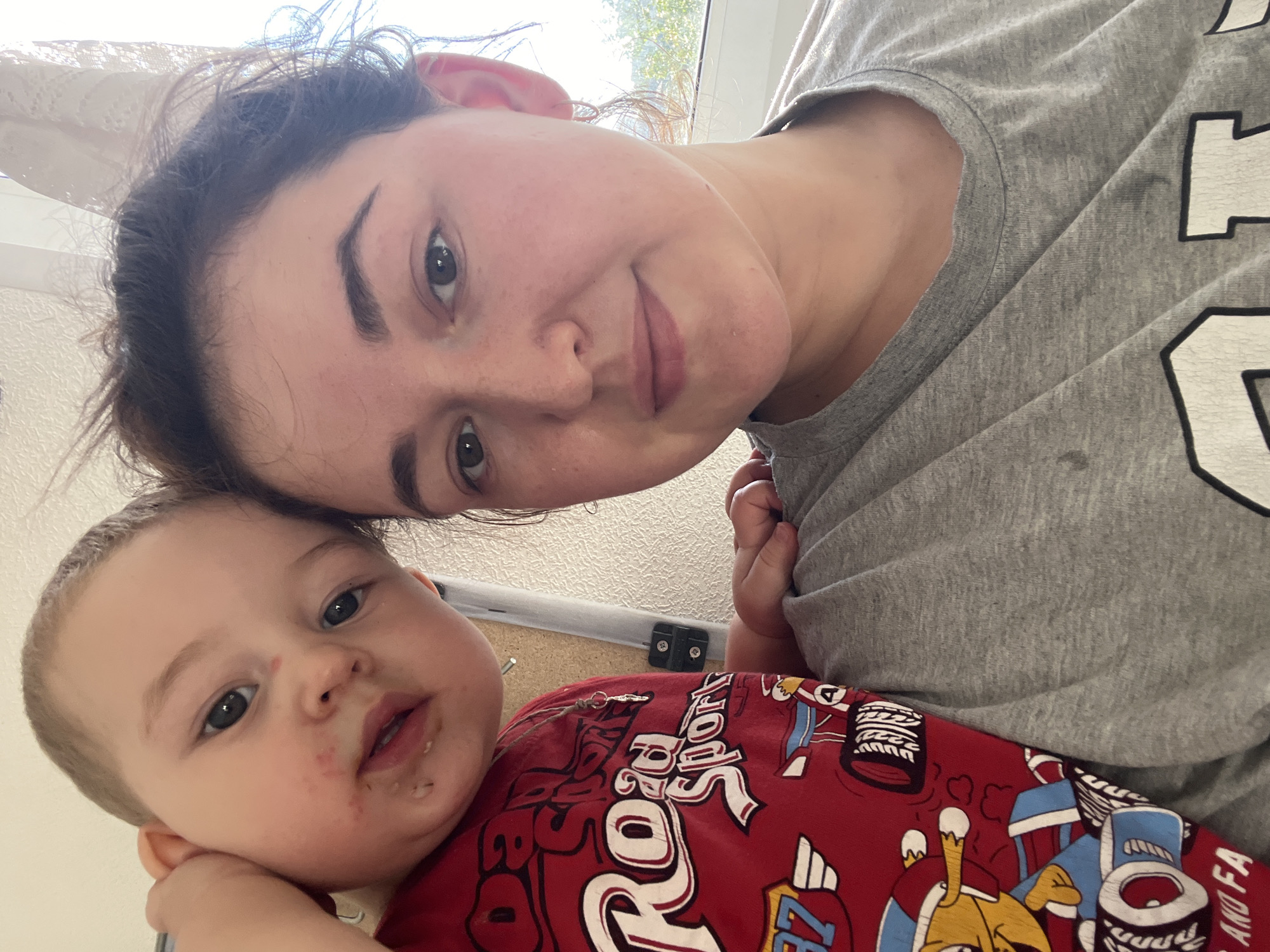This is a selfie of a woman and a baby. The woman is in the foreground, her face tilted towards the camera and slightly out of focus. She has dark hair and is wearing a grey t-shirt. To her left, and slightly lower, is a baby. The baby has light brown hair and big, dark eyes. The baby's face has some redness and what appears to be food residue around its mouth. The baby is wearing a red shirt with a graphic print of what looks like cartoon characters and text. The visible text on the baby's shirt reads "Roaring Sport".
The setting appears to be indoors, with a window in the background suggesting daylight. The overall lighting is bright, indicating it's daytime. There are no explicit cues to suggest the weather or a specific location within Chelyabinsk, Russia, beyond the provided context. The image conveys a sense of intimacy and a casual moment between the woman and the baby. The woman's expression is soft, and the baby looks directly at the camera.