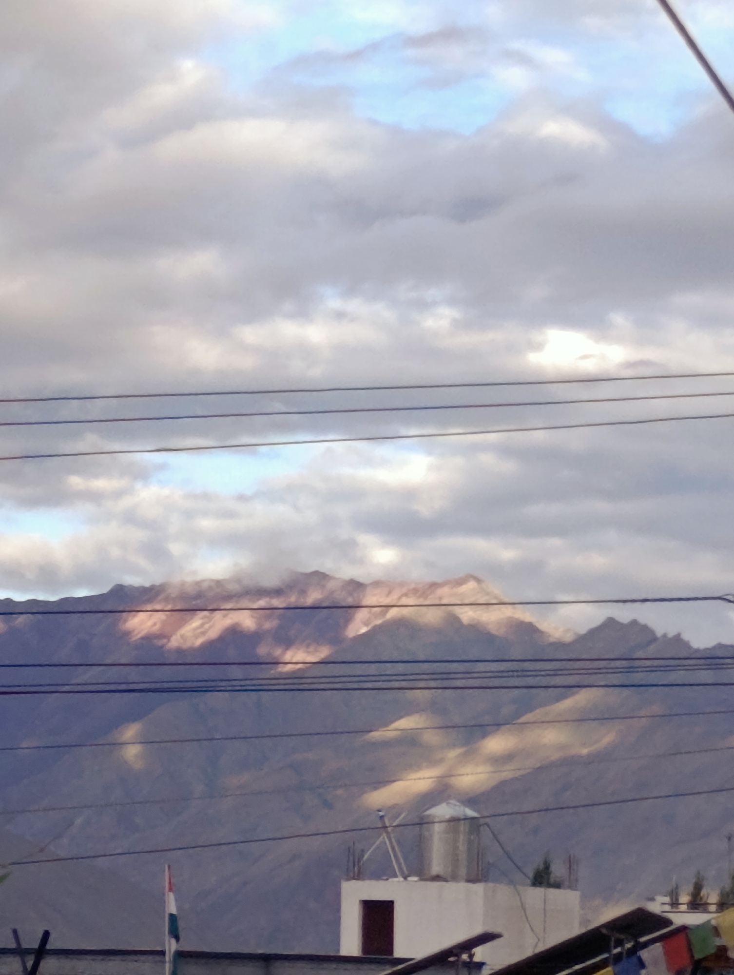 The image shows a mountainous landscape with a cloudy sky over the city of Leh, India. In the foreground, there are several power lines stretching across the frame. Below the power lines, a white building with a water tank on top is visible, along with part of a smaller building and some colorful flags. A flag with three horizontal stripes (saffron, white, and green) is planted in front of the white building. The background is dominated by rugged mountains, with sunlight illuminating some of their peaks and slopes, creating a contrast with the shadowed areas. The sky is filled with a mixture of white and gray clouds, with patches of blue sky visible in between. The overall scene suggests an urban area at the foothills of a mountain range, likely taken during the daytime.