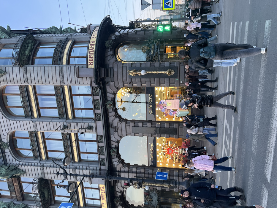 The image presents a street-level view of the historic Singer House (Dom Knigi), also known as the House of Books, situated at the intersection of Nevsky Prospekt and the Griboyedov Canal in Saint Petersburg, Russia. The multi-story building displays an ornate Art Nouveau architectural style with a grey facade, featuring numerous arched windows and decorative balconies. The ground floor is occupied by "Cafe René," identified by multiple illuminated signs within its large display windows, which showcase merchandise including a prominent red decorative wheel. A sign bearing "КОМПАНІЯ ЗИНГЕРЪ" (Kompania Zinger) is affixed to the building's facade, with "ДОМ КНИГИ" (Dom Knigi) partially visible near the corner entrance.
A bustling pedestrian scene unfolds in the foreground and on the sidewalk. Numerous individuals are seen walking and congregating, dressed in varied cool-weather attire such as jackets, coats, and hoodies. On the white-striped crosswalk, a person in a black jacket carrying a bottle walks left-to-right, a person in a pink puffer jacket walks right-to-left, and a person in a black puffer jacket looking at a mobile phone walks left-to-right. A person viewed from behind, wearing a black leather jacket and a teal backpack, walks away from the camera on the crosswalk. To the right, a pedestrian traffic light displays a green walking figure and the numerical countdown "20." A street lamp and a blue directional sign are visible on the left, while overhead wires traverse the light blue sky.