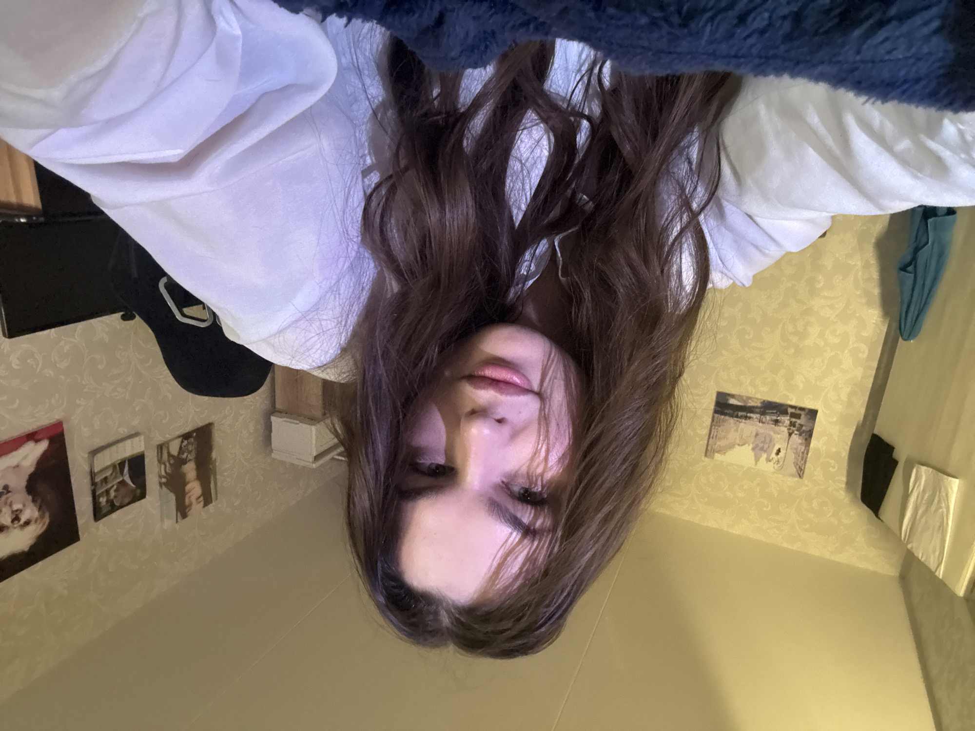 A photo taken upside down, presumably by the person in the image. The subject is a fair-skinned individual with brown hair, wearing a white shirt. The background appears to be a room with patterned wallpaper. There are pictures on the wall. Location is unclear. No action or interaction is taking place in the image. The photo was likely taken indoors. The city is unidentifiable based on the image.