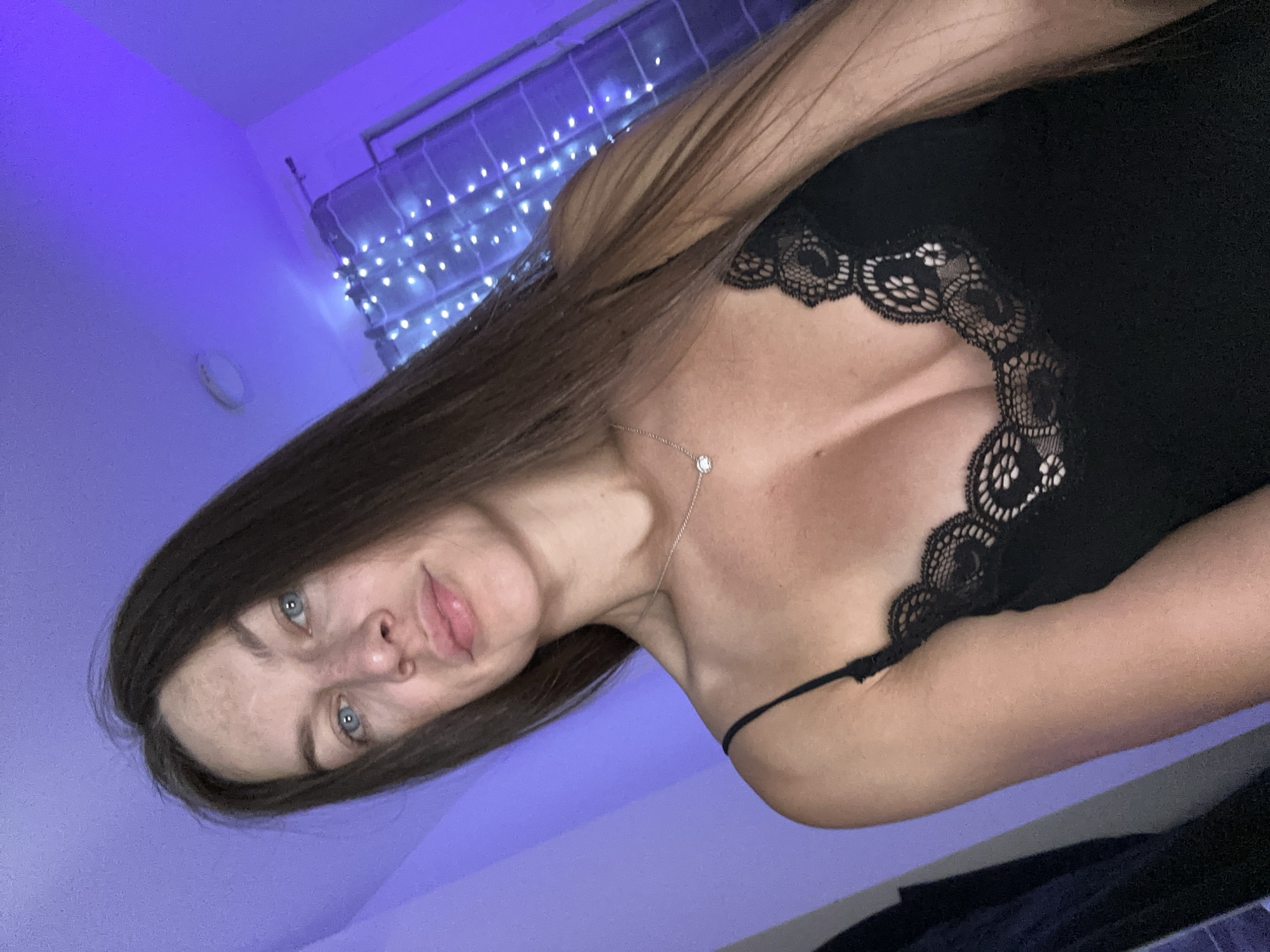 A young woman with long brown hair and blue eyes is shown in a selfie. She is wearing a black camisole with lace trim and a delicate silver necklace with a small pendant. The background is dimly lit with purple ambient light, and a string of fairy lights is visible, suggesting an indoor setting. The woman's expression is neutral as she looks directly at the camera. The overall atmosphere is intimate and casual. There is no information about the specific location in Donji Pobori, Montenegro.